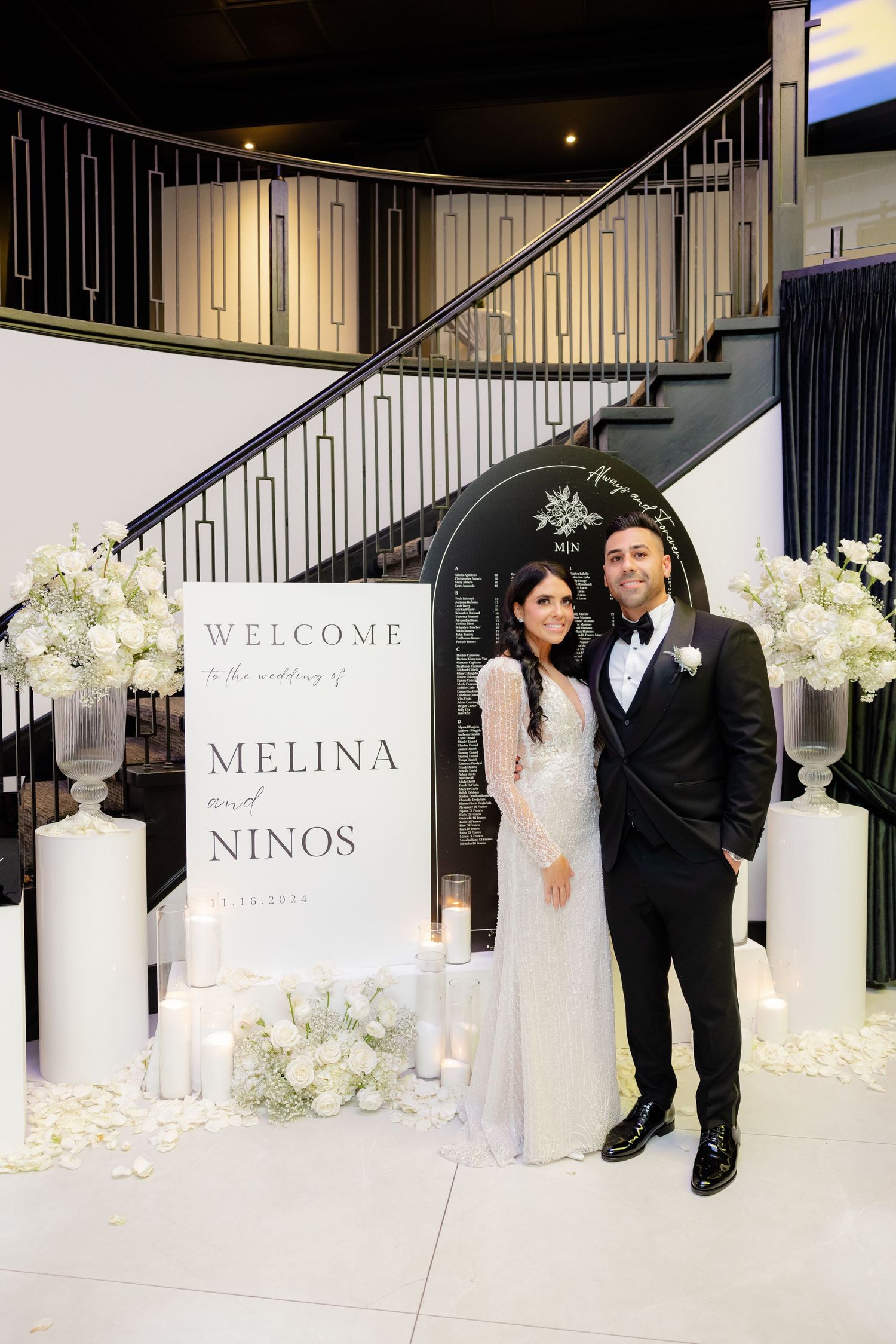Elegant wedding venue entrance with white floral arrangements and welcome sign against staircase backdrop.