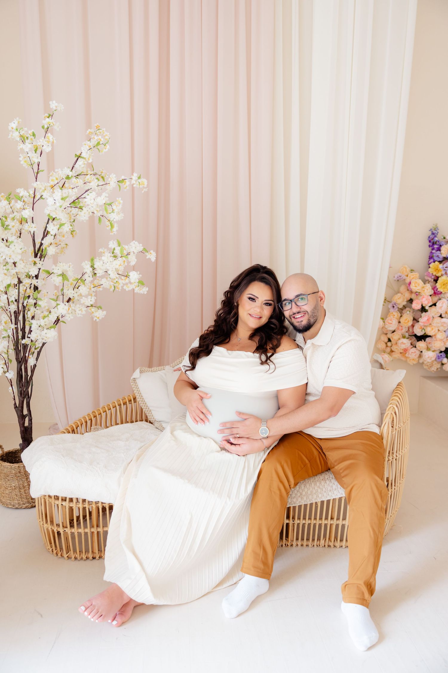 A couple poses together on a rattan chair with floral decorations and soft blush draping in the background.