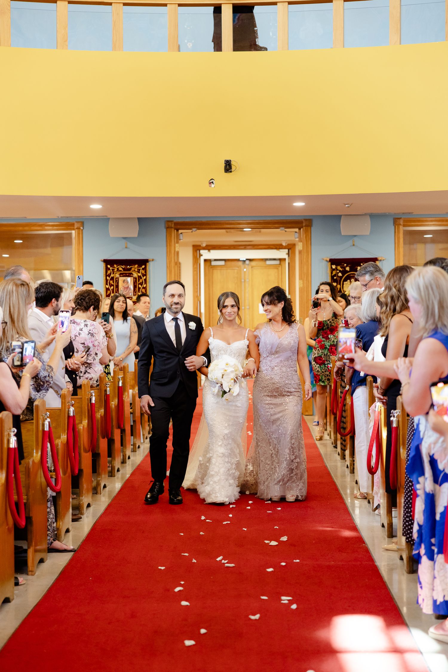 Wedding processional walking down red-carpeted church aisle with guests watching from wooden pews.