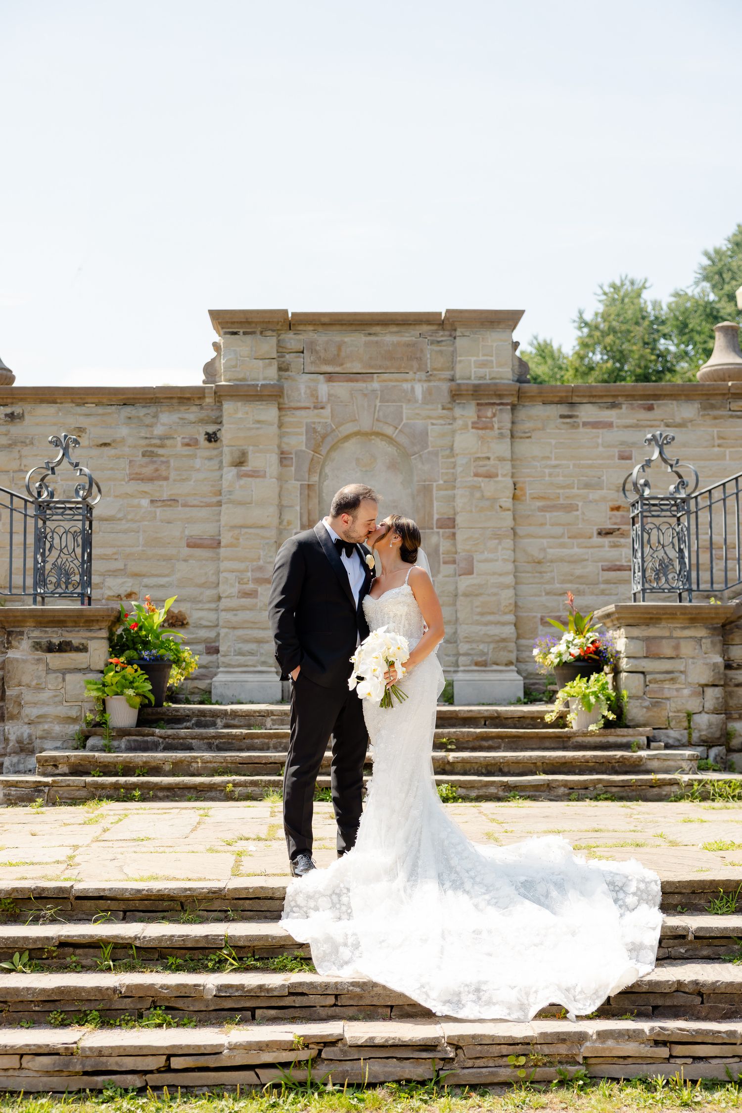 A romantic wedding scene on stone steps with ornate iron gates and beautiful floral arrangements at an elegant venue.