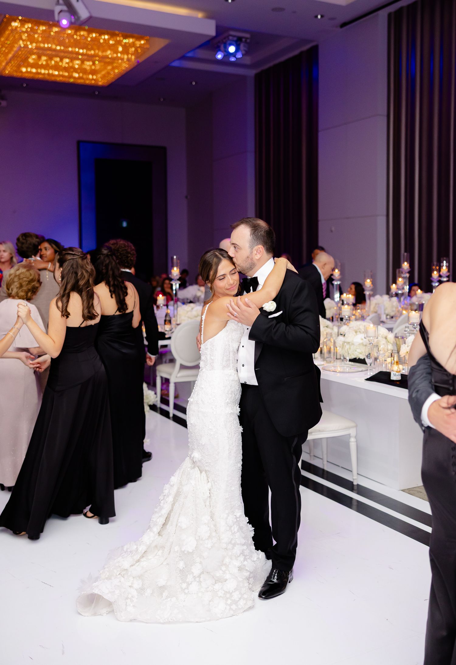 A couple shares a romantic dance at their elegant wedding reception with white decor and purple lighting.