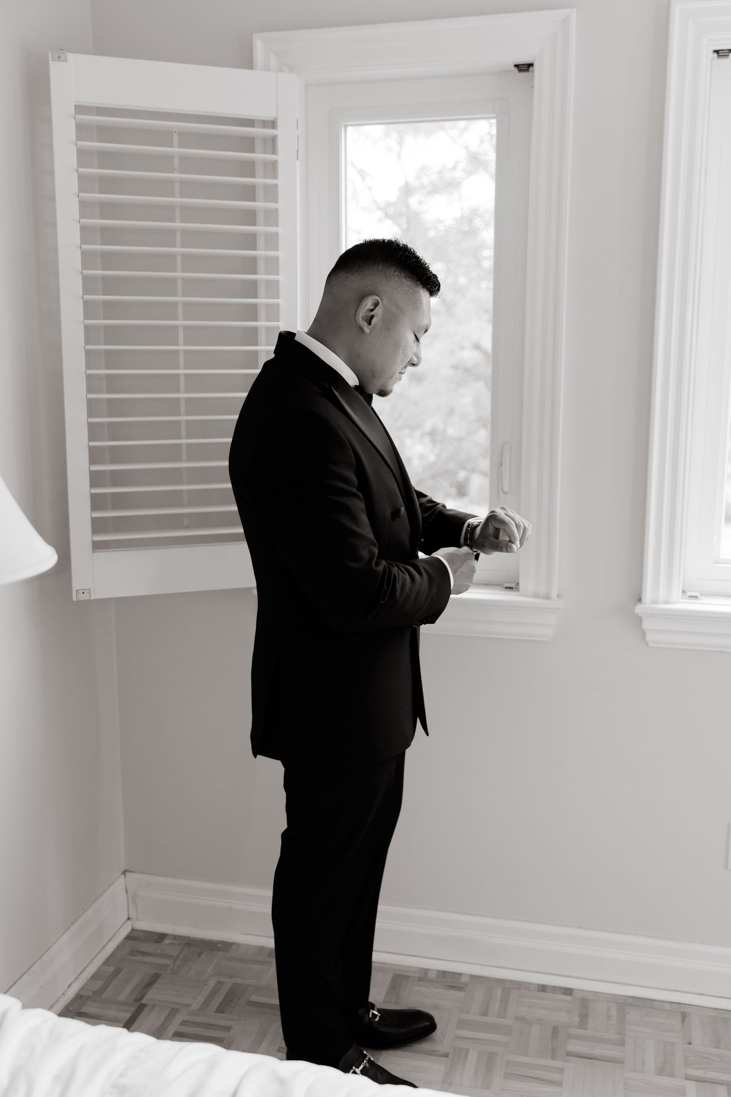Man in black business suit inspects white window shutters in a bright room with hardwood flooring.