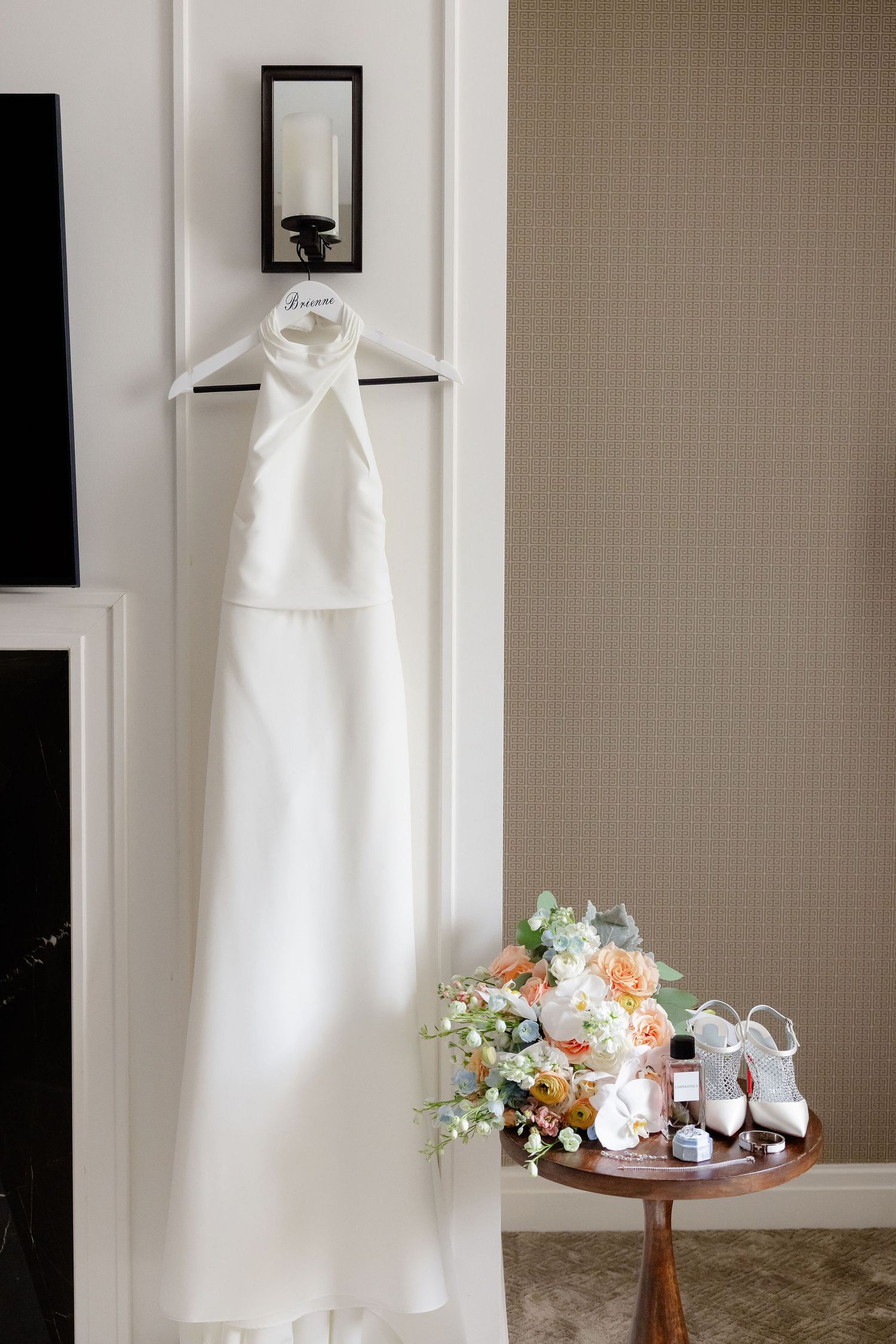 A white wedding dress hangs elegantly next to floral arrangements and decor on a wooden table in a sunlit room.