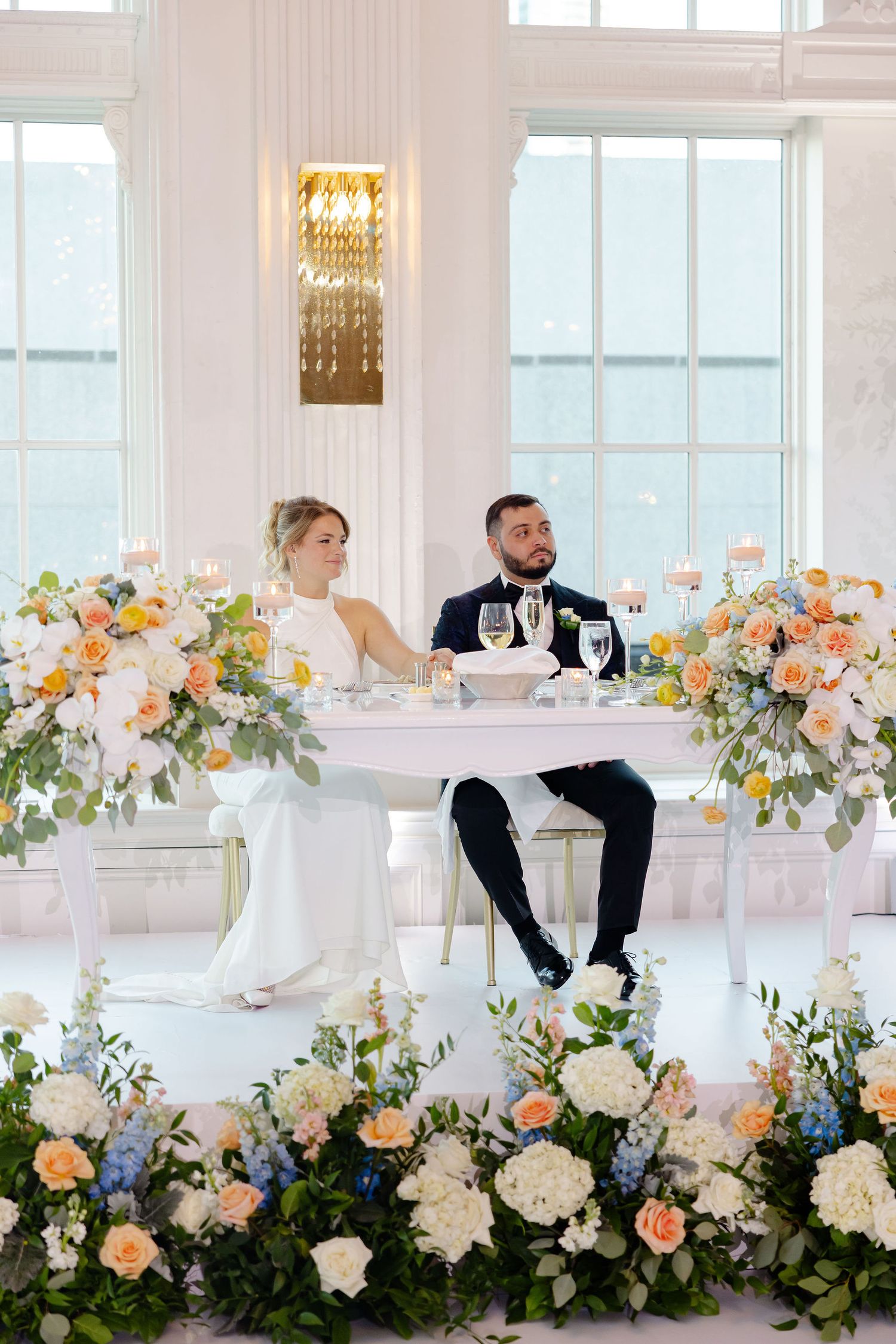 Couple sitting at elegant wedding reception table adorned with lush peach and white floral arrangements and greenery.