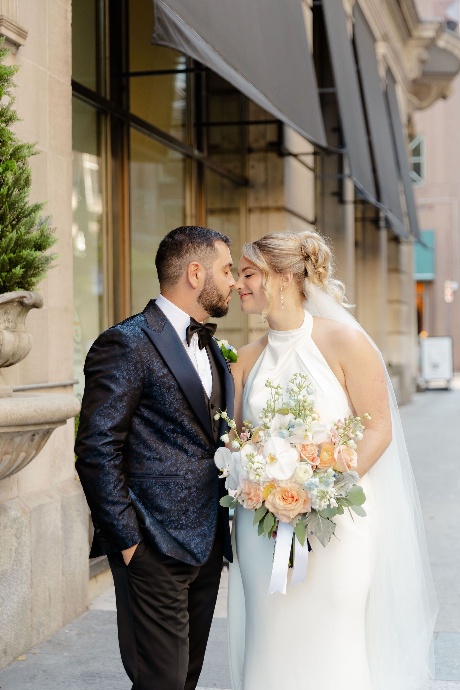 A romantic wedding moment captured outside a building with elegant attire and peach floral bouquet.