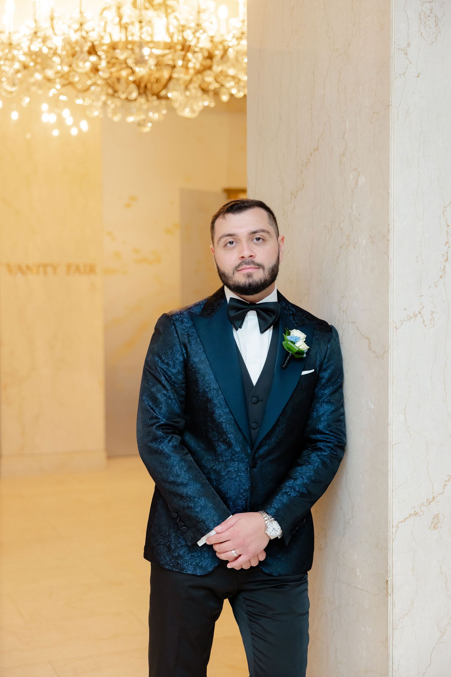 Formal portraits of groom in black tuxedo with boutonniere under elegant chandelier lighting.