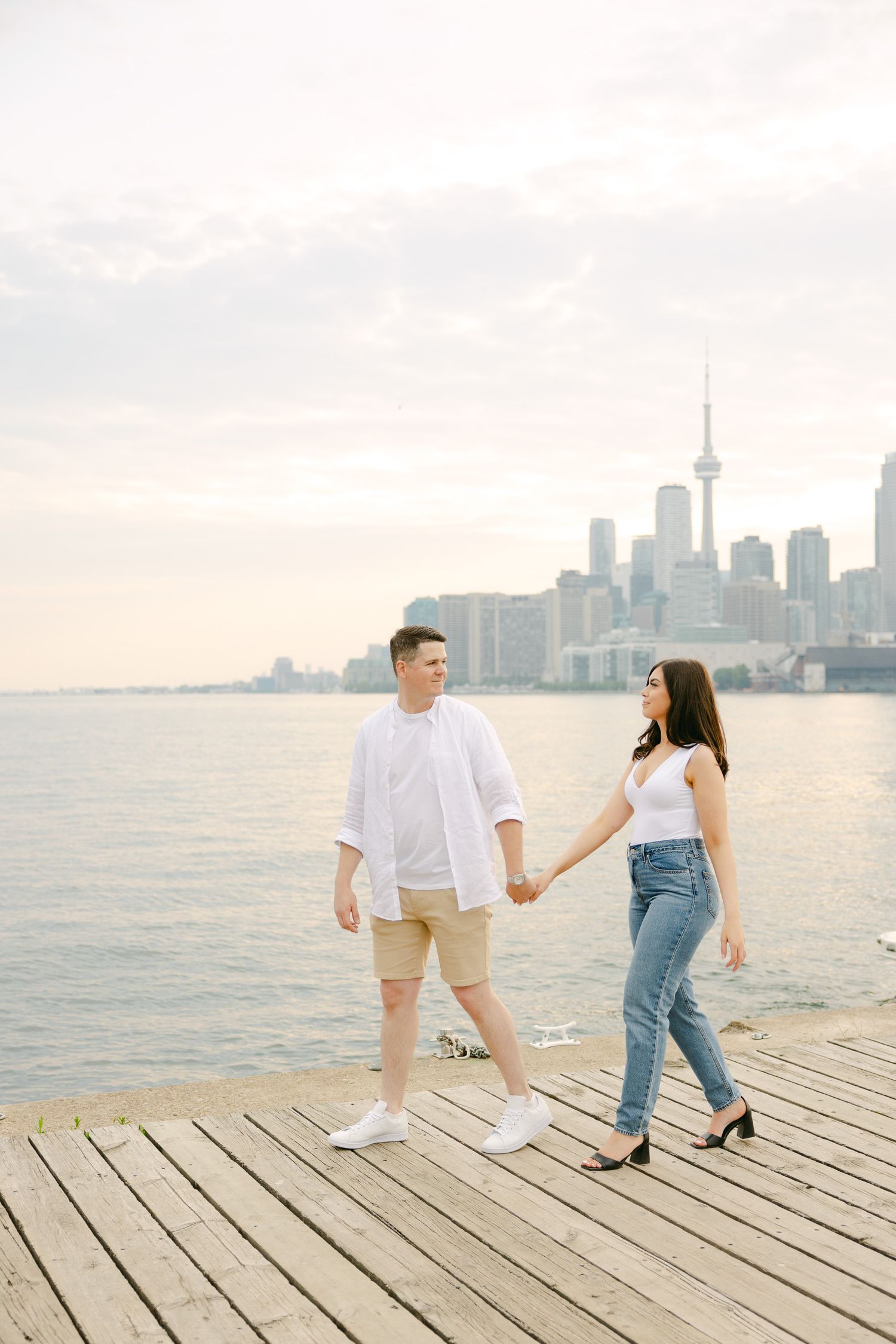 Couple walking hand in hand on wooden boardwalk with Toronto waterfront view.