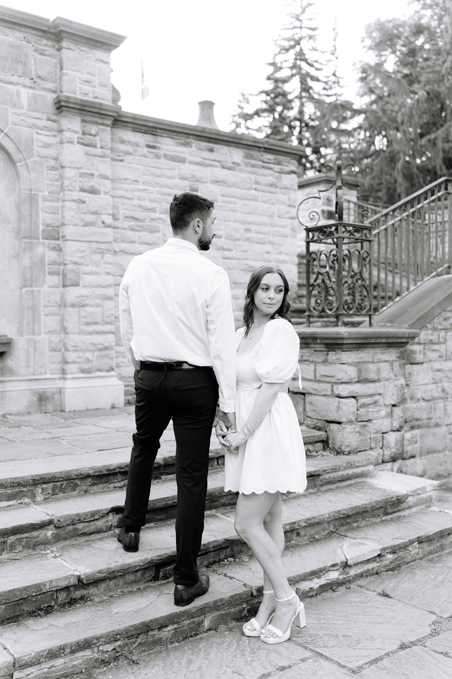 A romantic couple stands on stone steps in front of a historic building with ornate wrought iron railings.