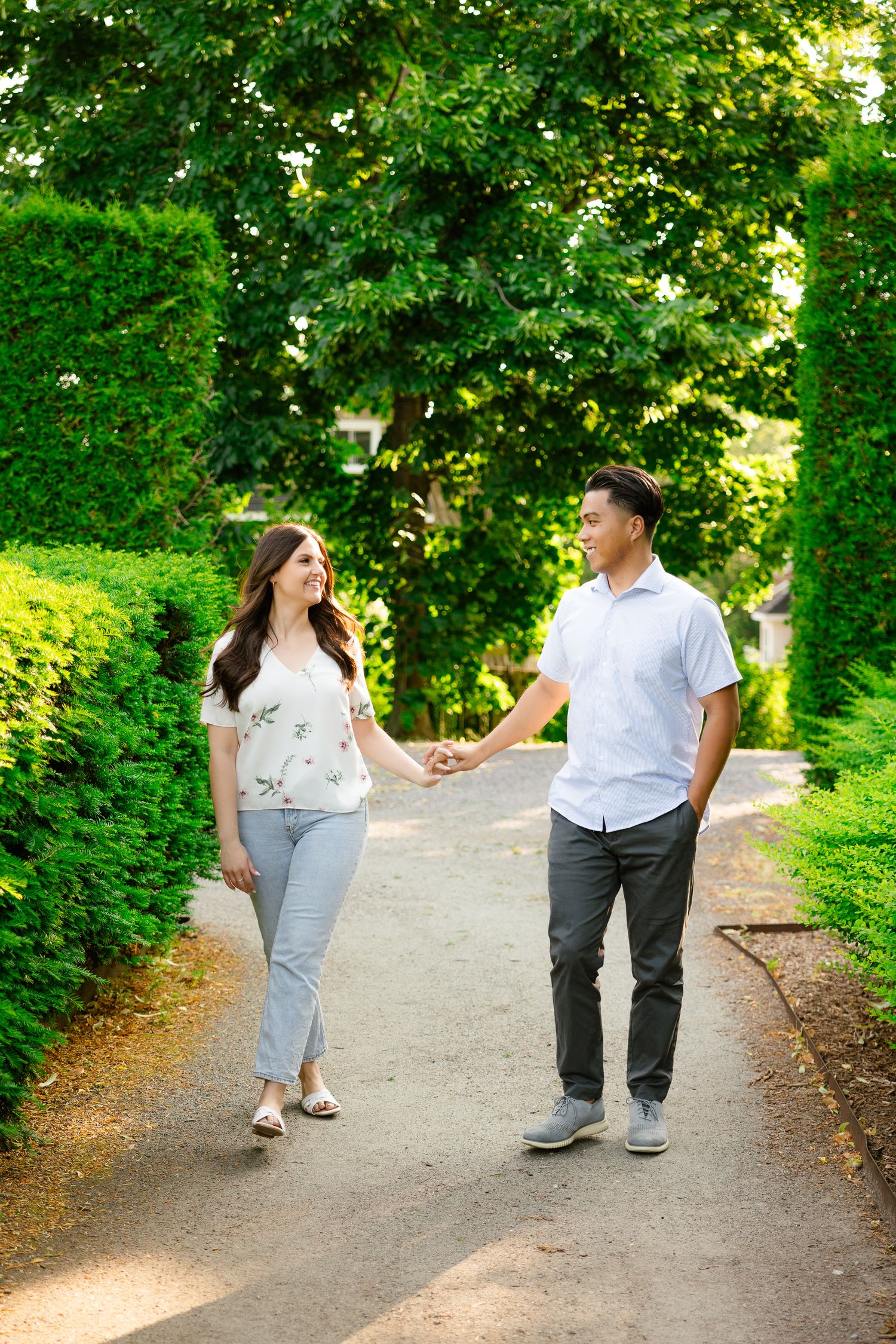 A couple holds hands while walking together on a garden path surrounded by green hedges.