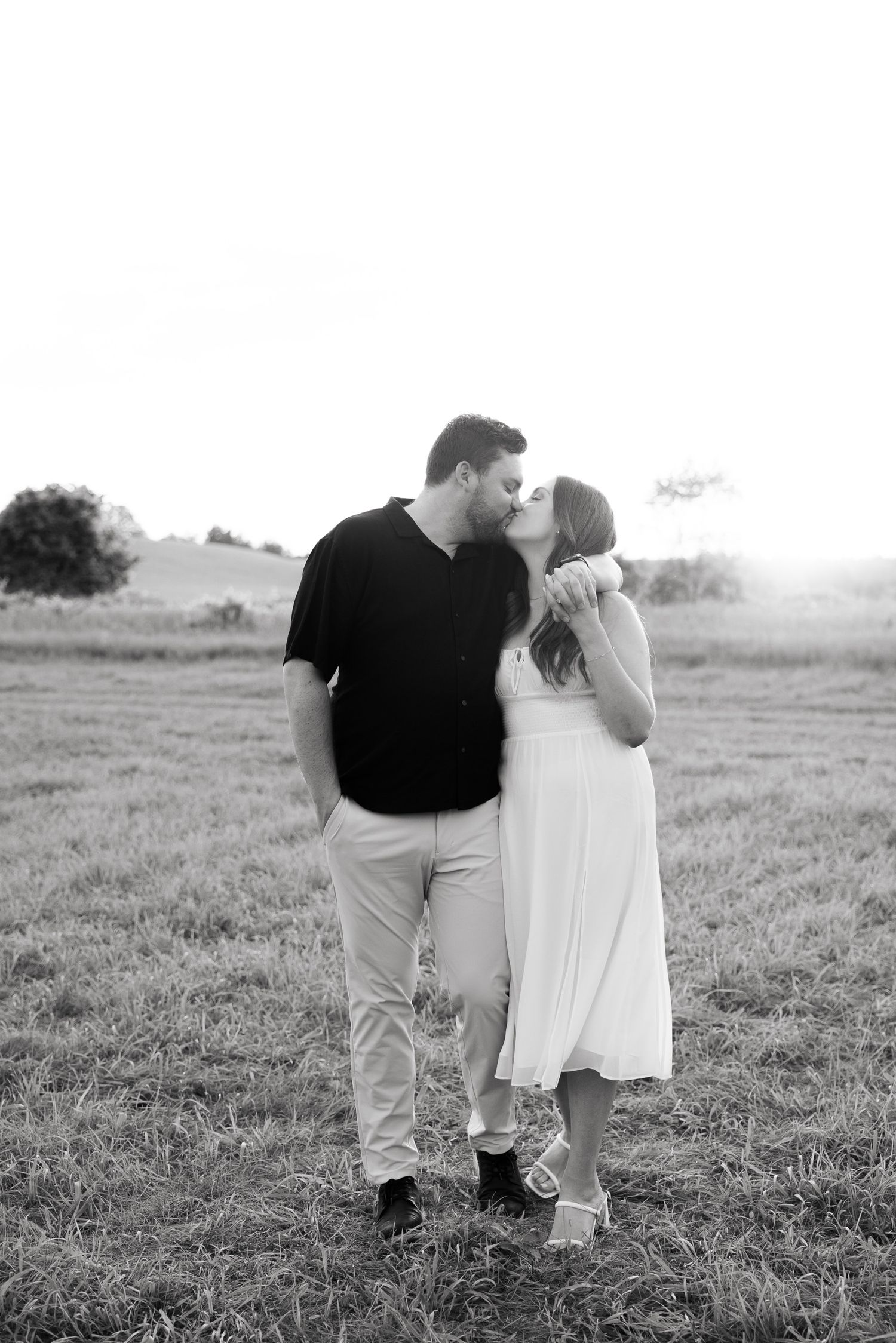 A romantic sunset engagement photo of a couple in a field with the person in black kissing their partner in white.