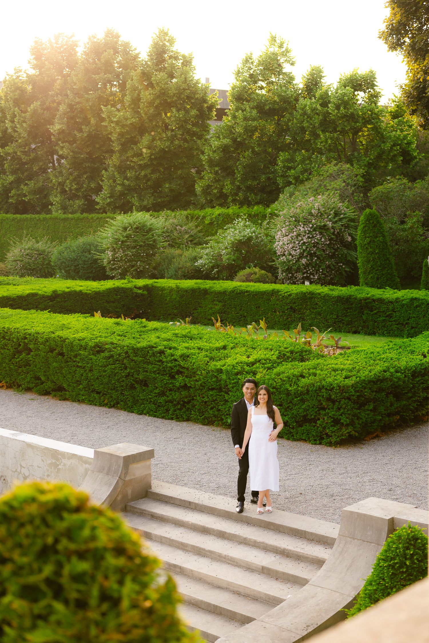 A couple standing on garden steps during sunset, surrounded by perfectly manicured hedges and trees.