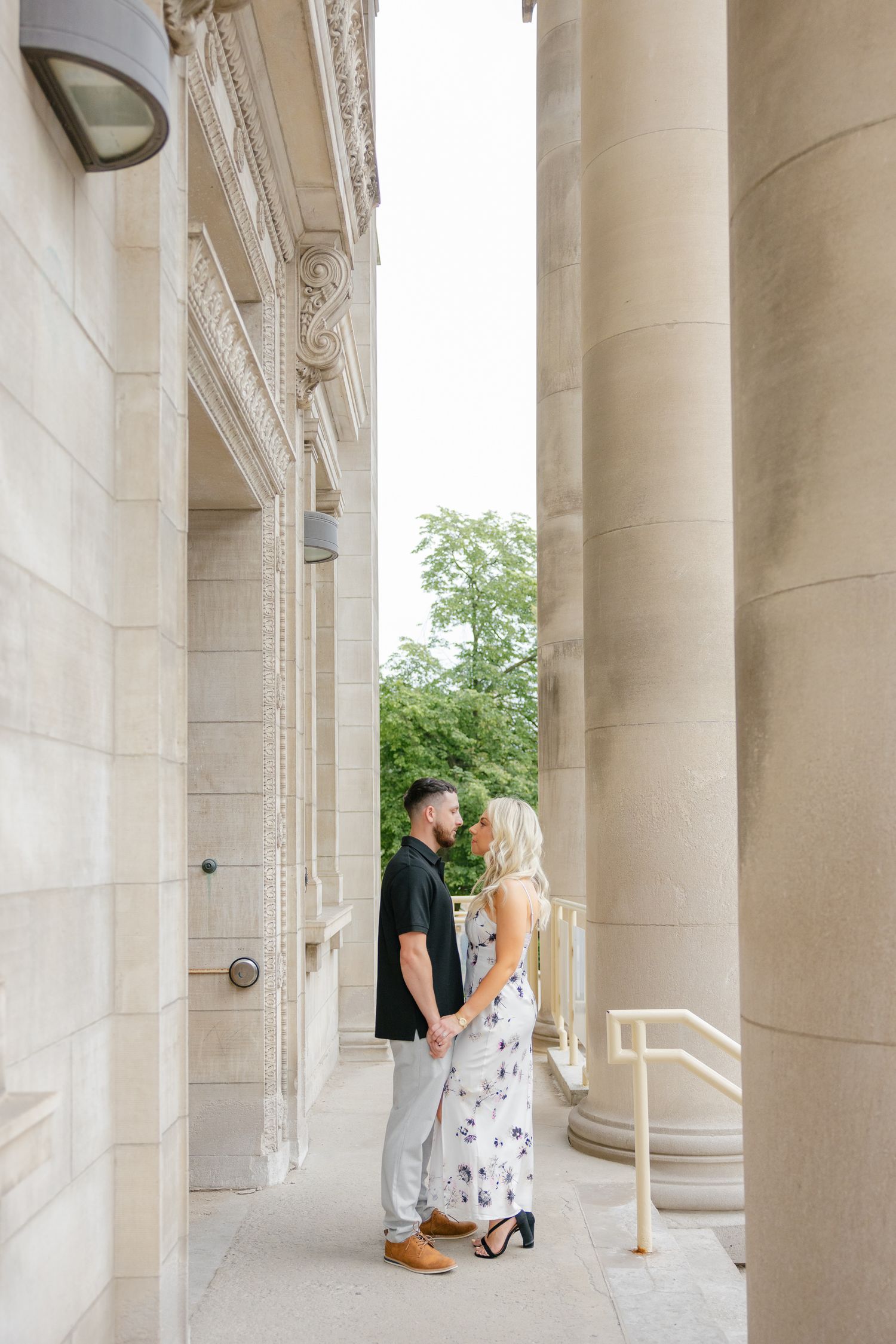 A couple poses romantically between elegant stone columns in formal attire on a bright summer day.
