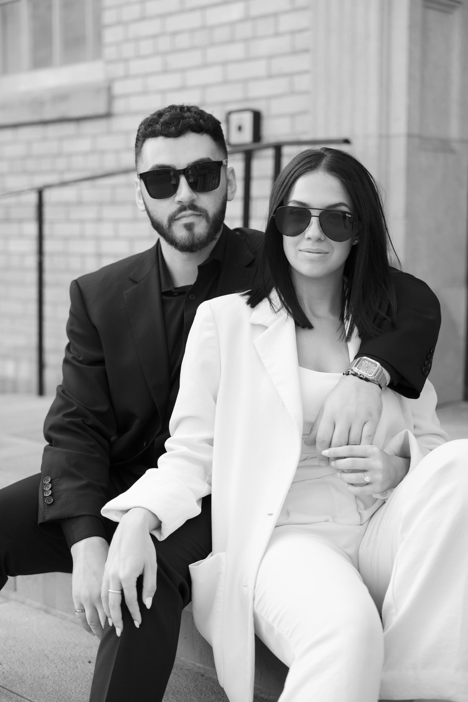 Monochrome portrait of stylish couple in sunglasses and formal wear sitting close together.