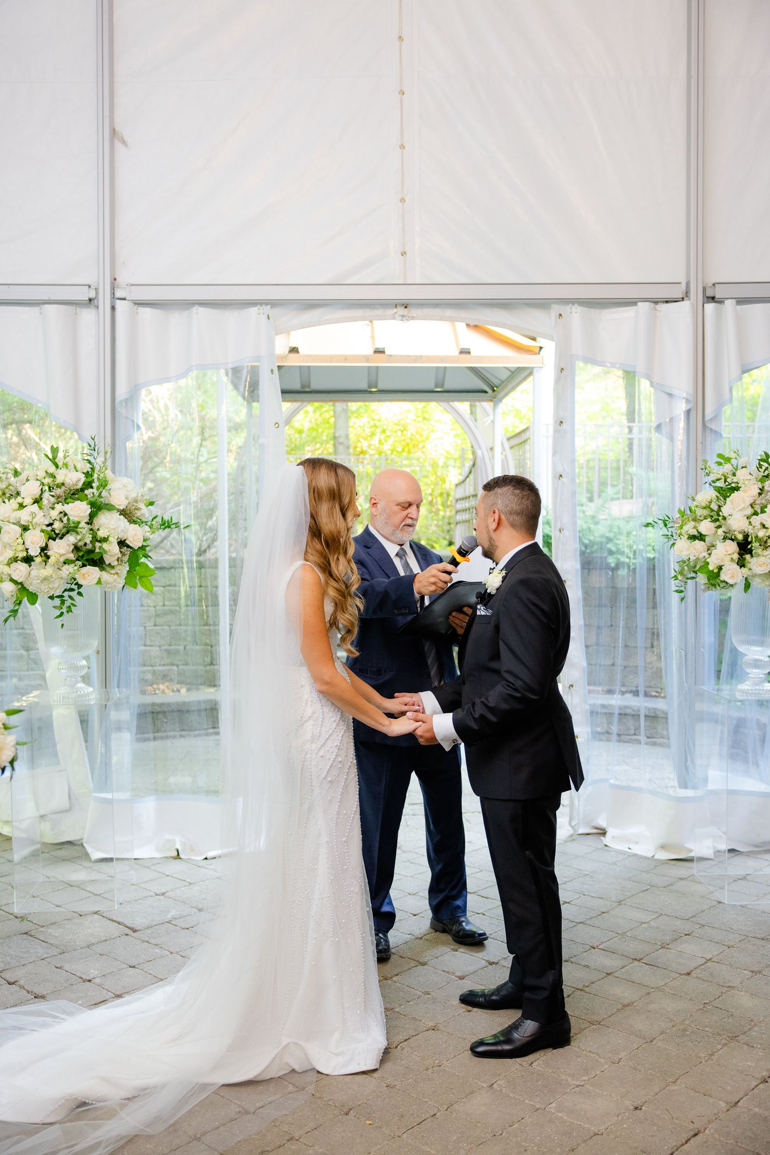 Wedding ceremony taking place under white tent with floral arrangements and stone flooring.