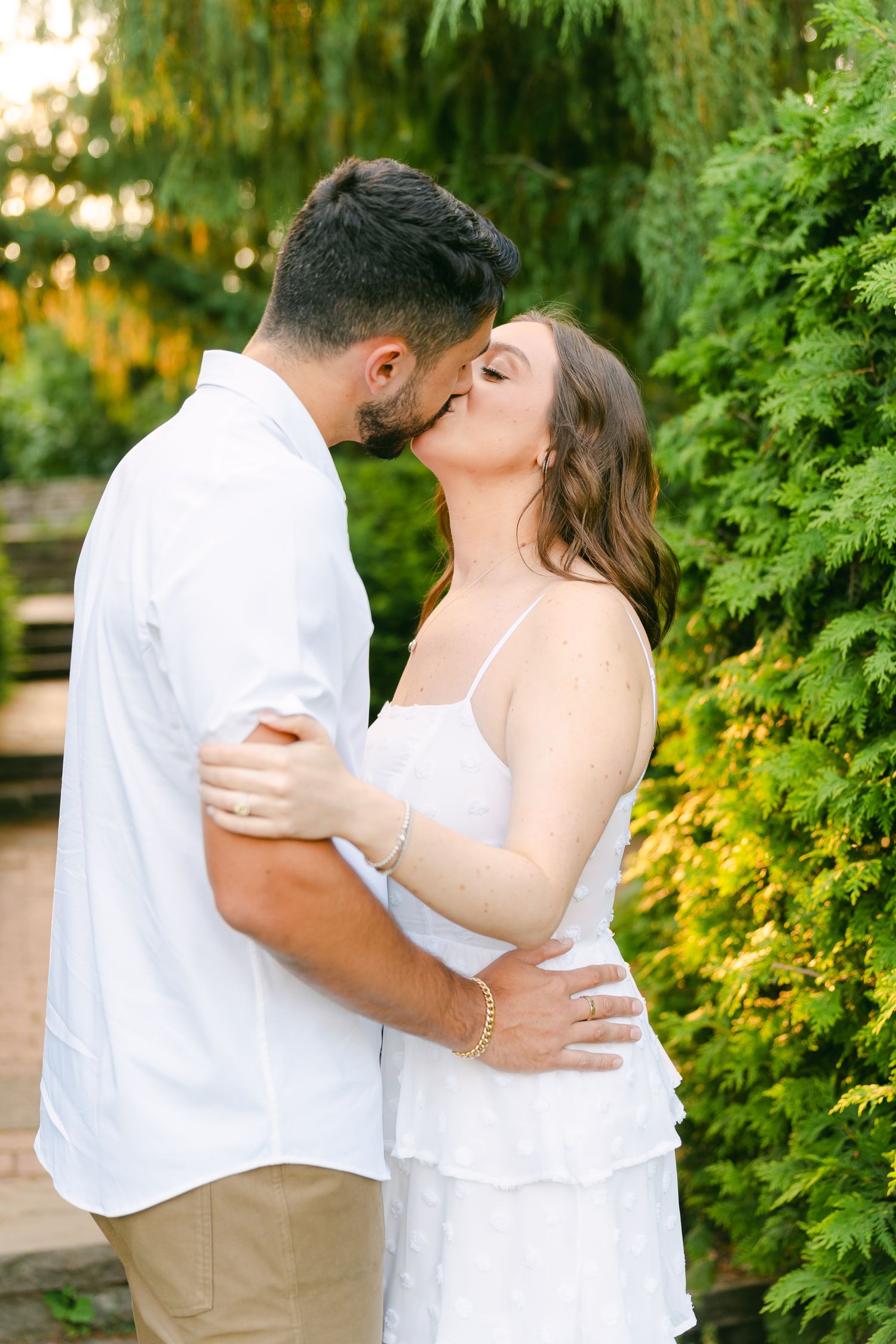 A romantic couple embraces and kisses in a garden setting with lush greenery and golden sunlight.