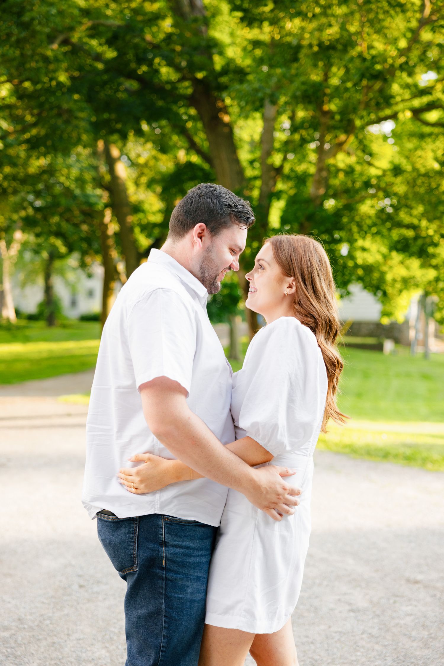 A romantic couple embracing on a tree-lined path in a sunlit park.