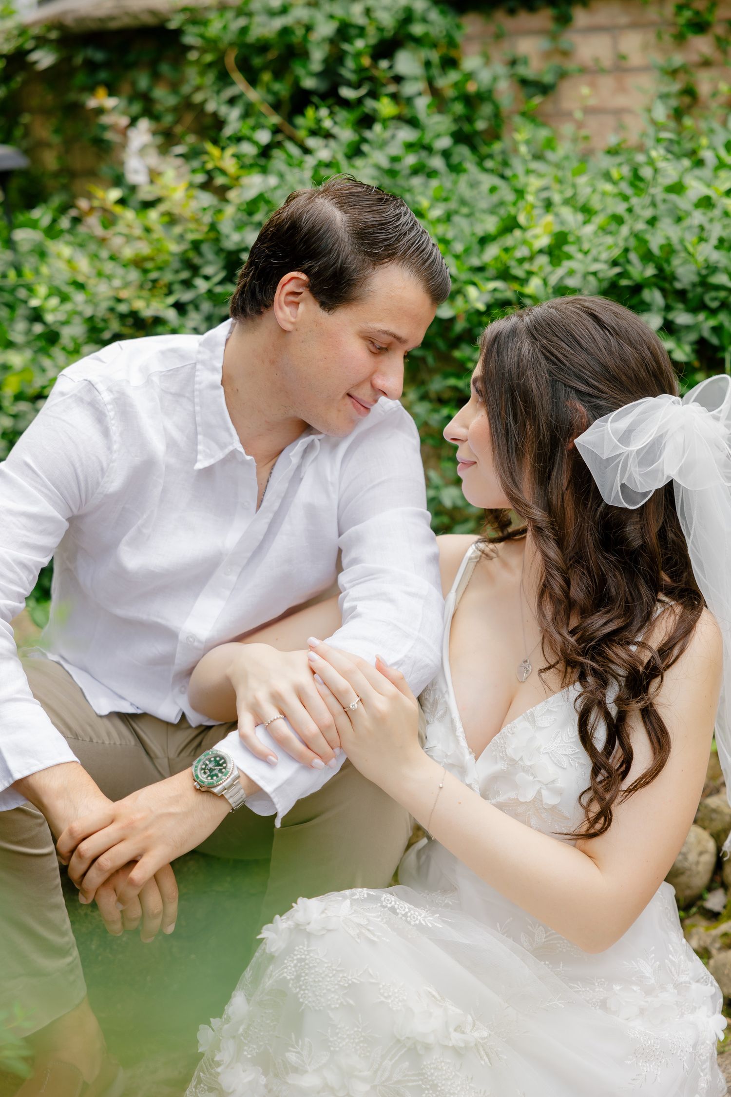 A tender moment between a couple sitting in a garden with brick walls and green foliage.