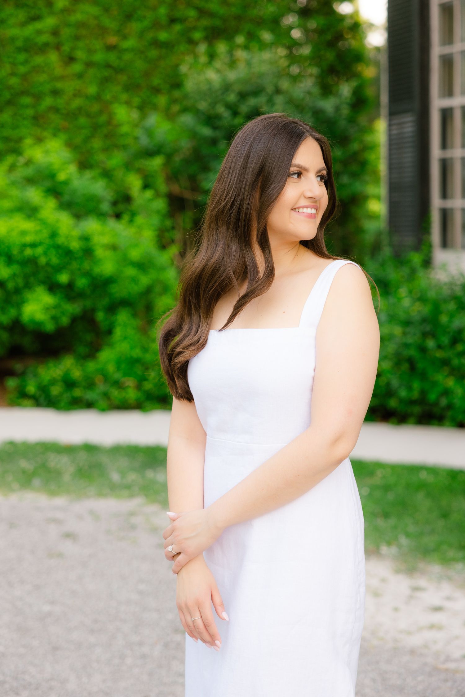 A woman in a white dress smiles while posing against a backdrop of spring greenery.