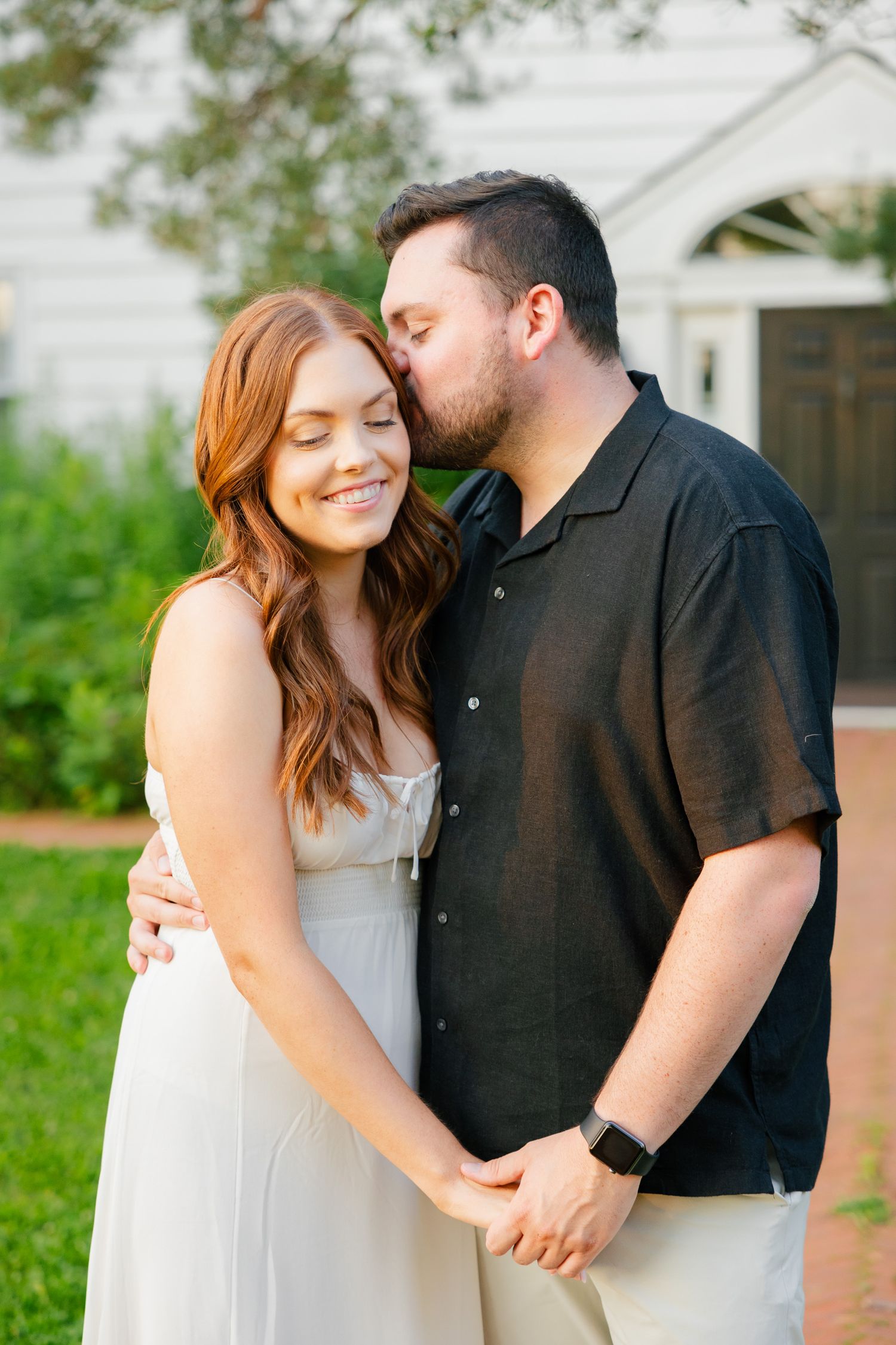 A sweet moment between a couple in front of a white house with green landscaping.