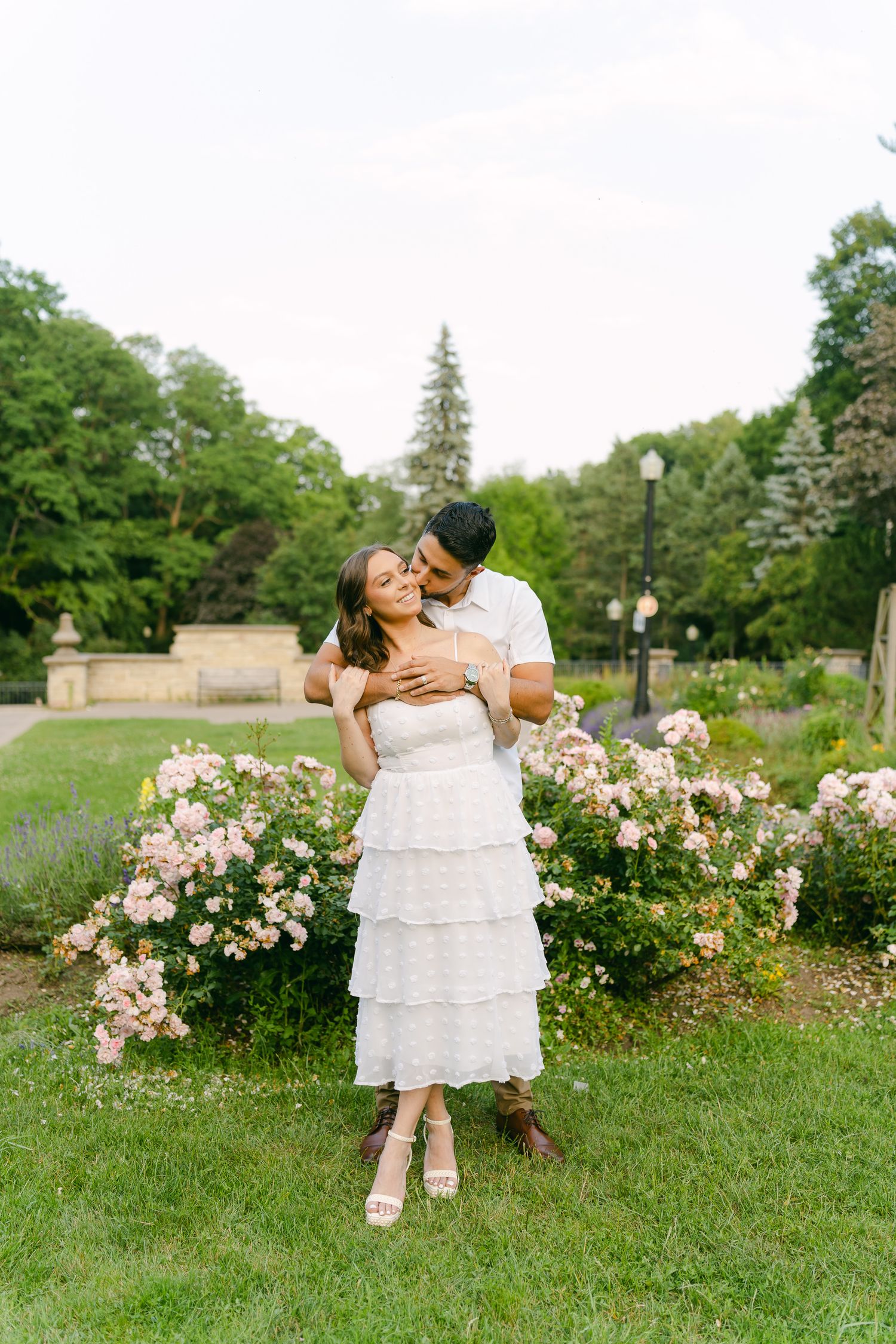 A joyful embrace in a formal garden with pink roses and evergreen trees in the background.