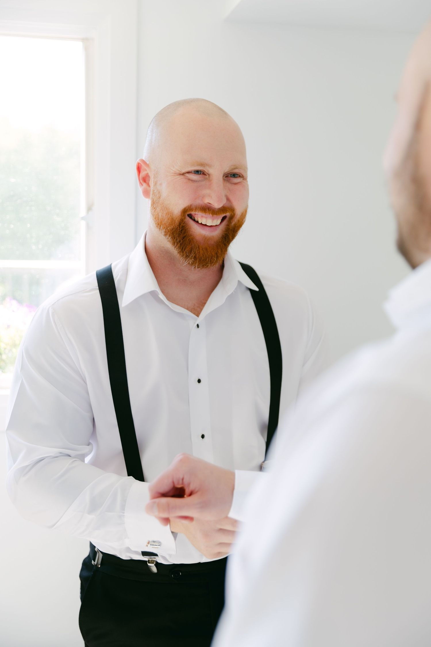 A person wearing white dress shirt and black suspenders smiling while getting ready.