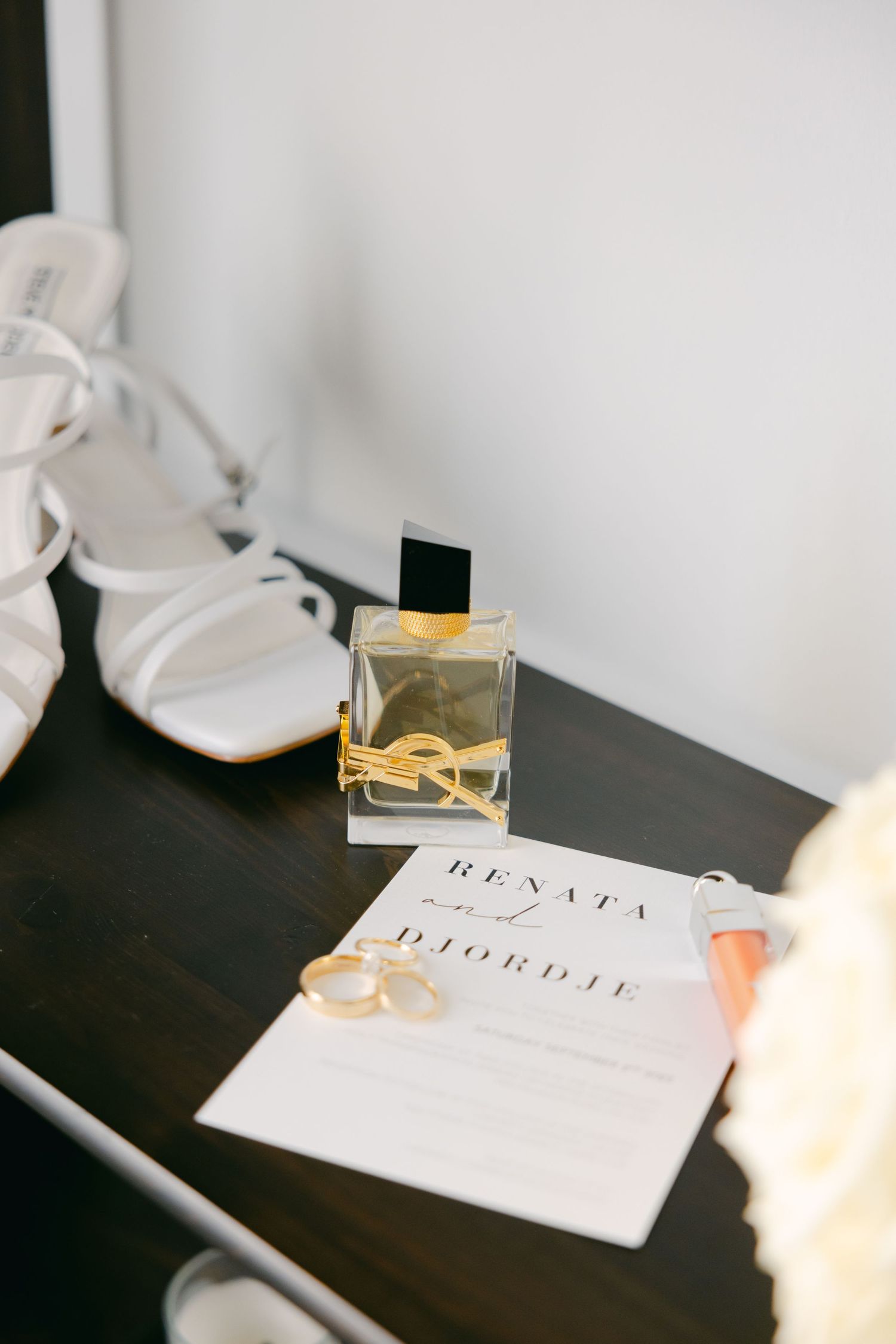 Wedding details including shoes, invitation, and perfume bottle arranged on dark surface.