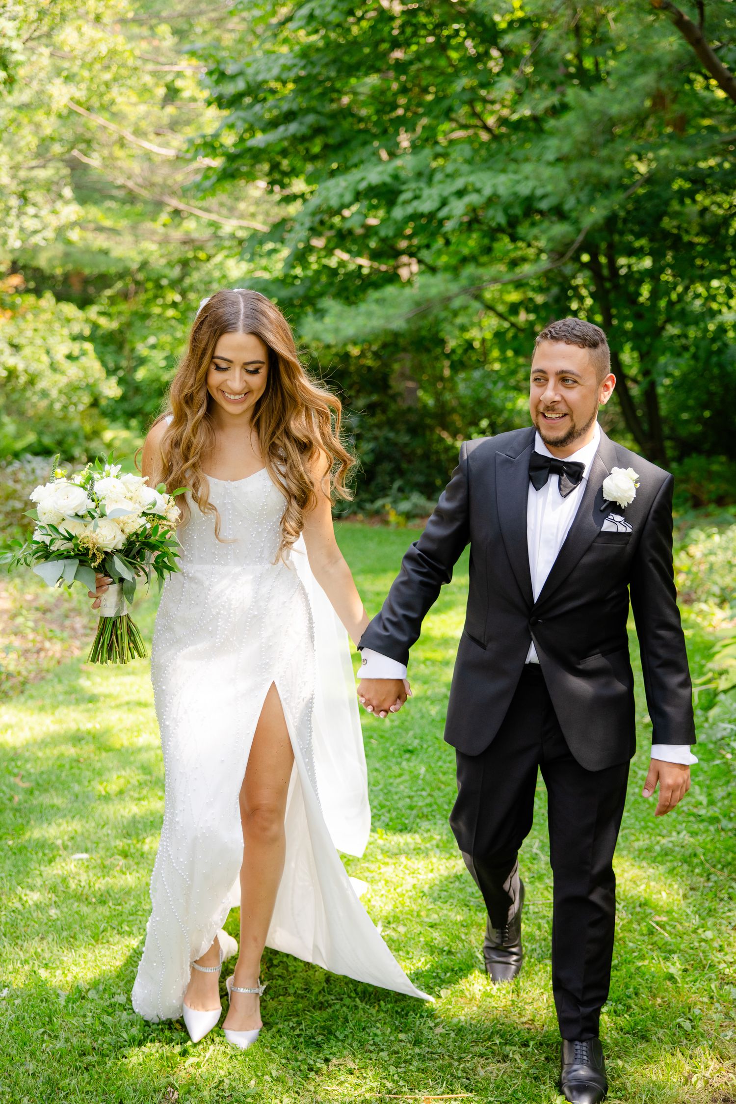 A wedding couple walks hand in hand on a lush lawn, with the bride in a slit white gown and groom in classic tuxedo.