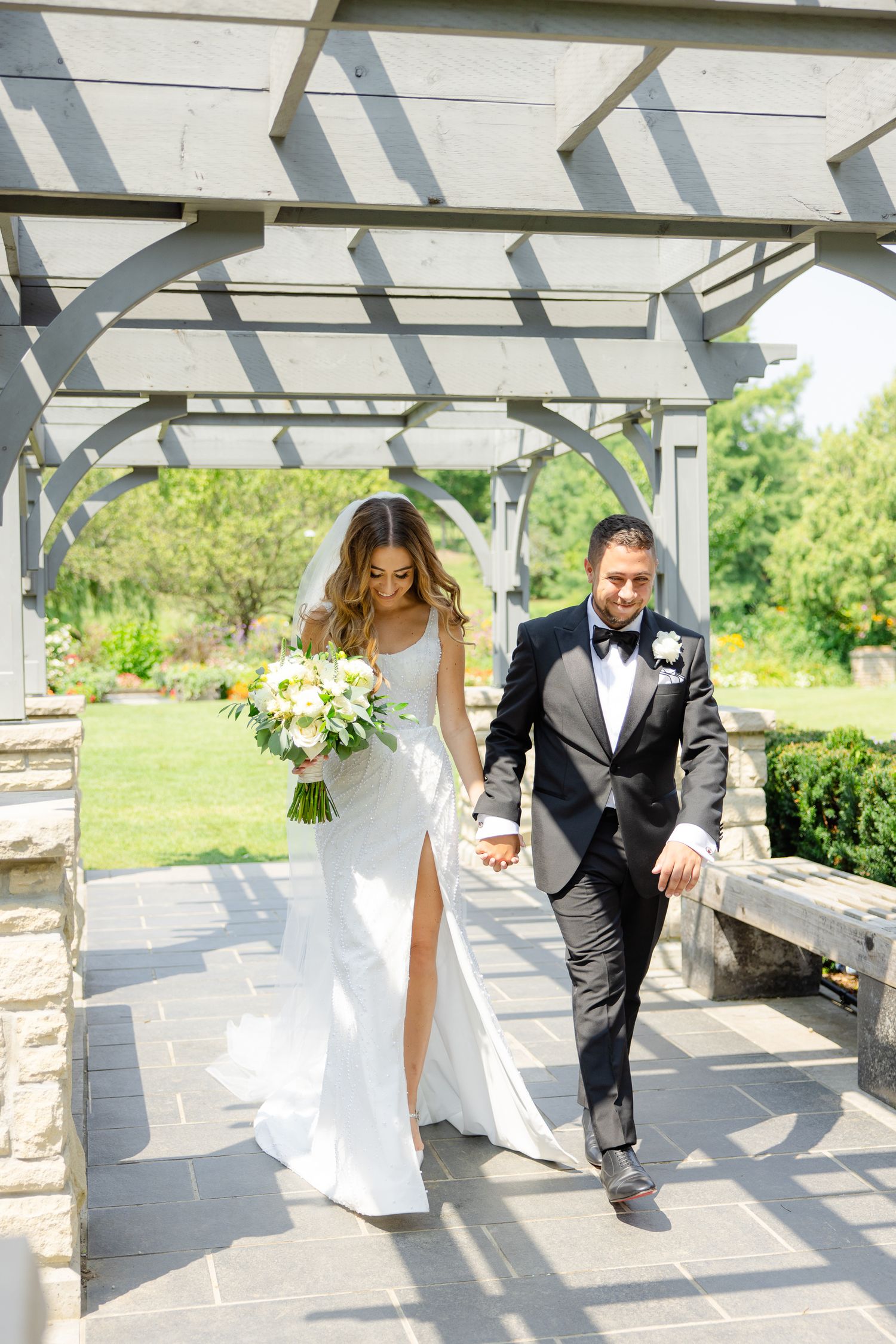 Wedding couple walking under an elegant white pergola with bride holding a white and green bouquet.