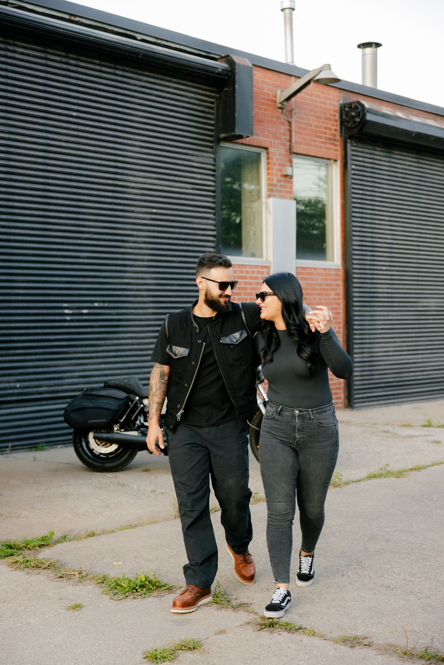 A stylish couple in black outfits walking together near a motorcycle and industrial building.