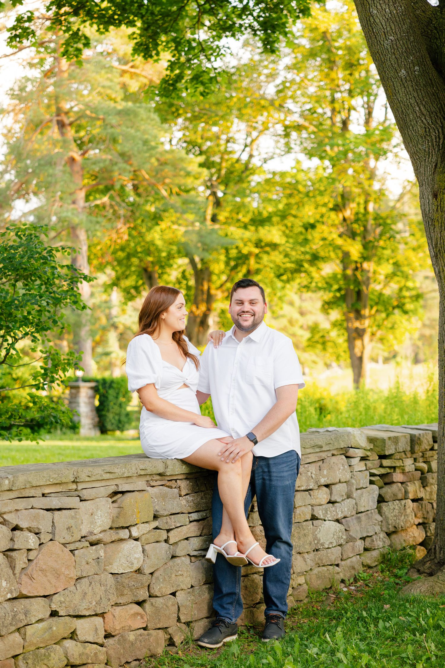 A couple shares a joyful moment together on a stone wall in a sunlit garden setting.