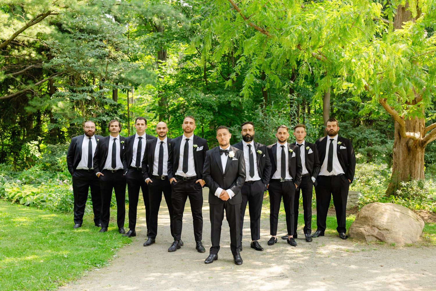 Groomsmen in matching black suits and ties stand in formation on a garden path.