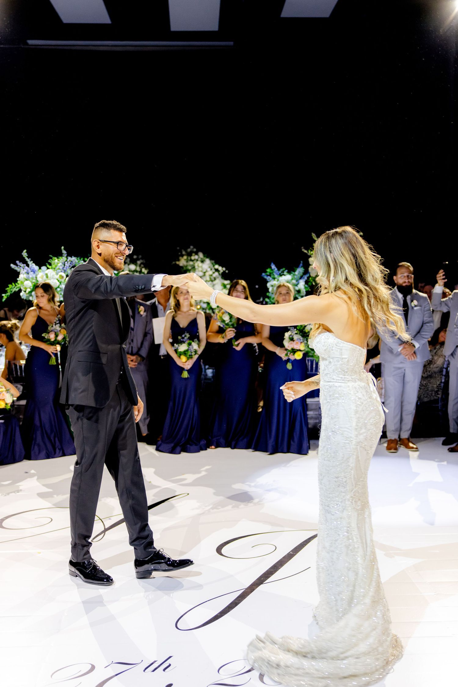 Newlyweds dancing at their reception with wedding party in navy and grey attire looking on.