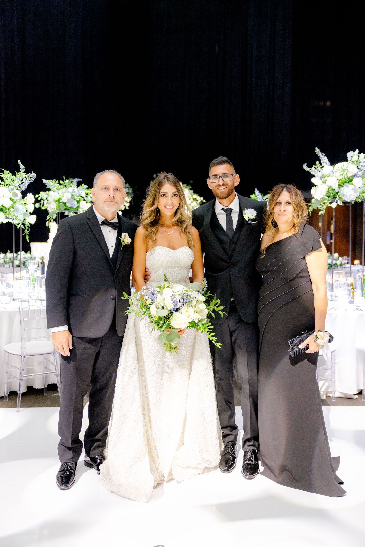 Elegant wedding party poses together in formal attire with white floral arrangements in the background.
