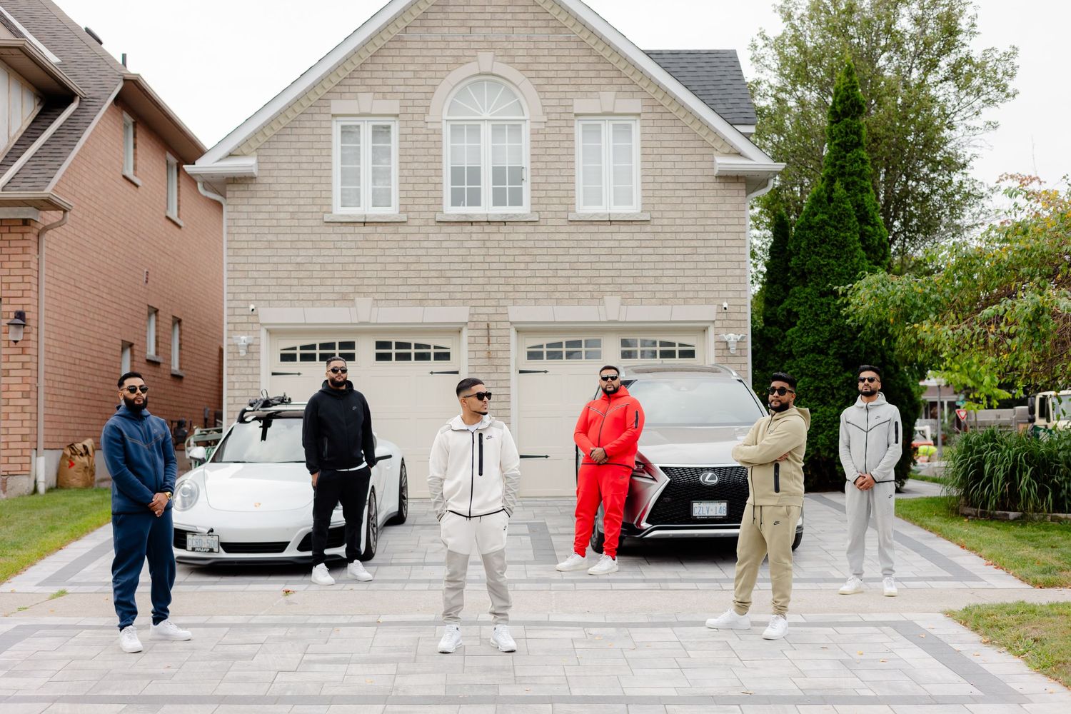 People in coordinated athleisure outfits stand on a driveway with luxury cars and a brick house backdrop.