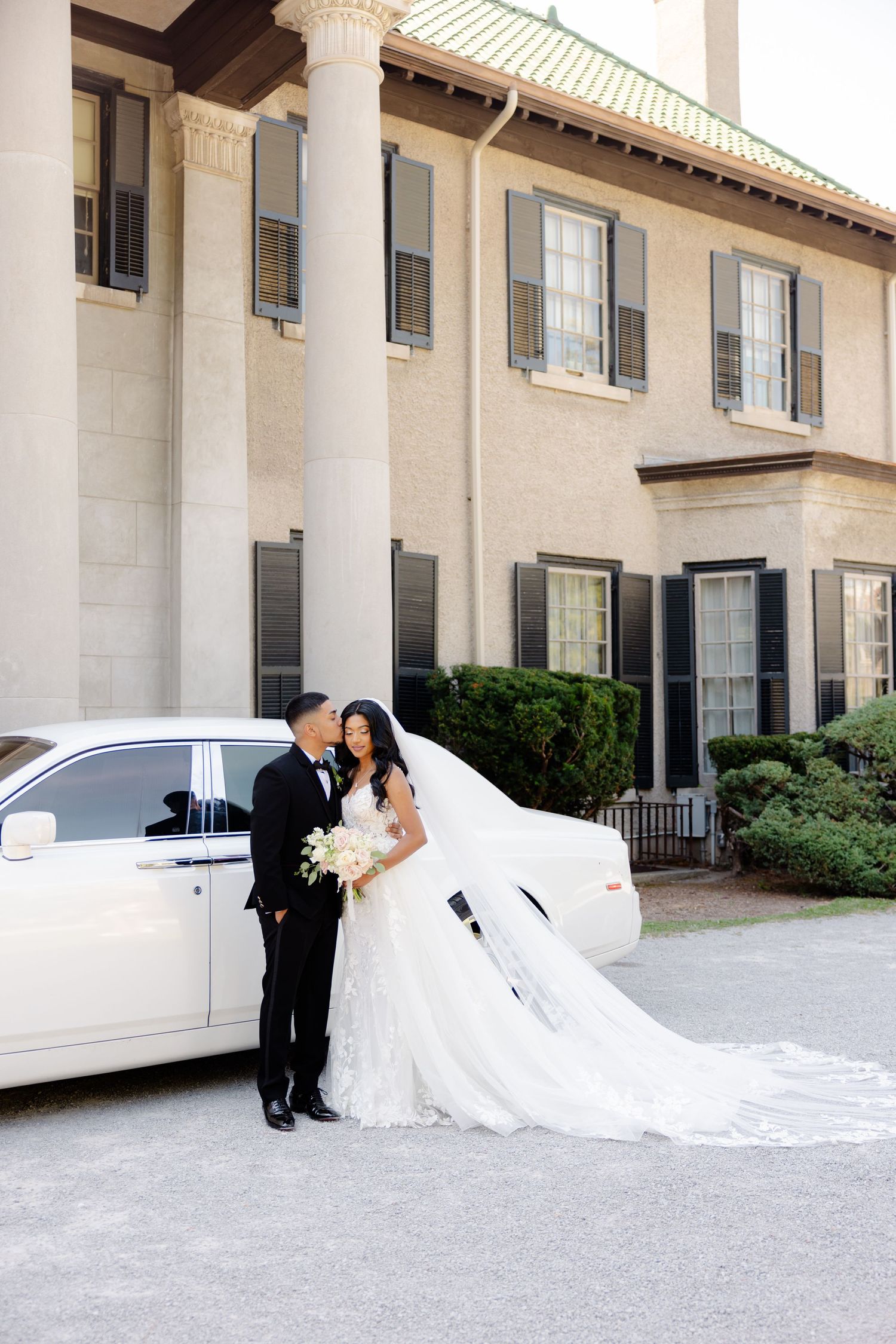 A newlywed couple stands beside a white luxury car with a flowing wedding dress train.