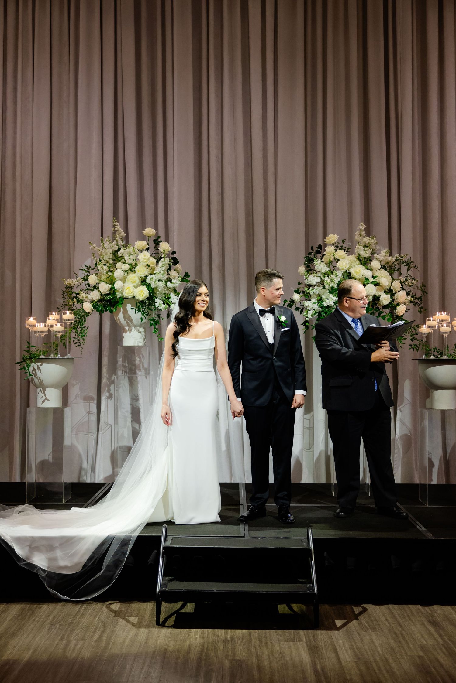 Modern wedding ceremony setup with luxurious floral displays and taupe draped backdrop.