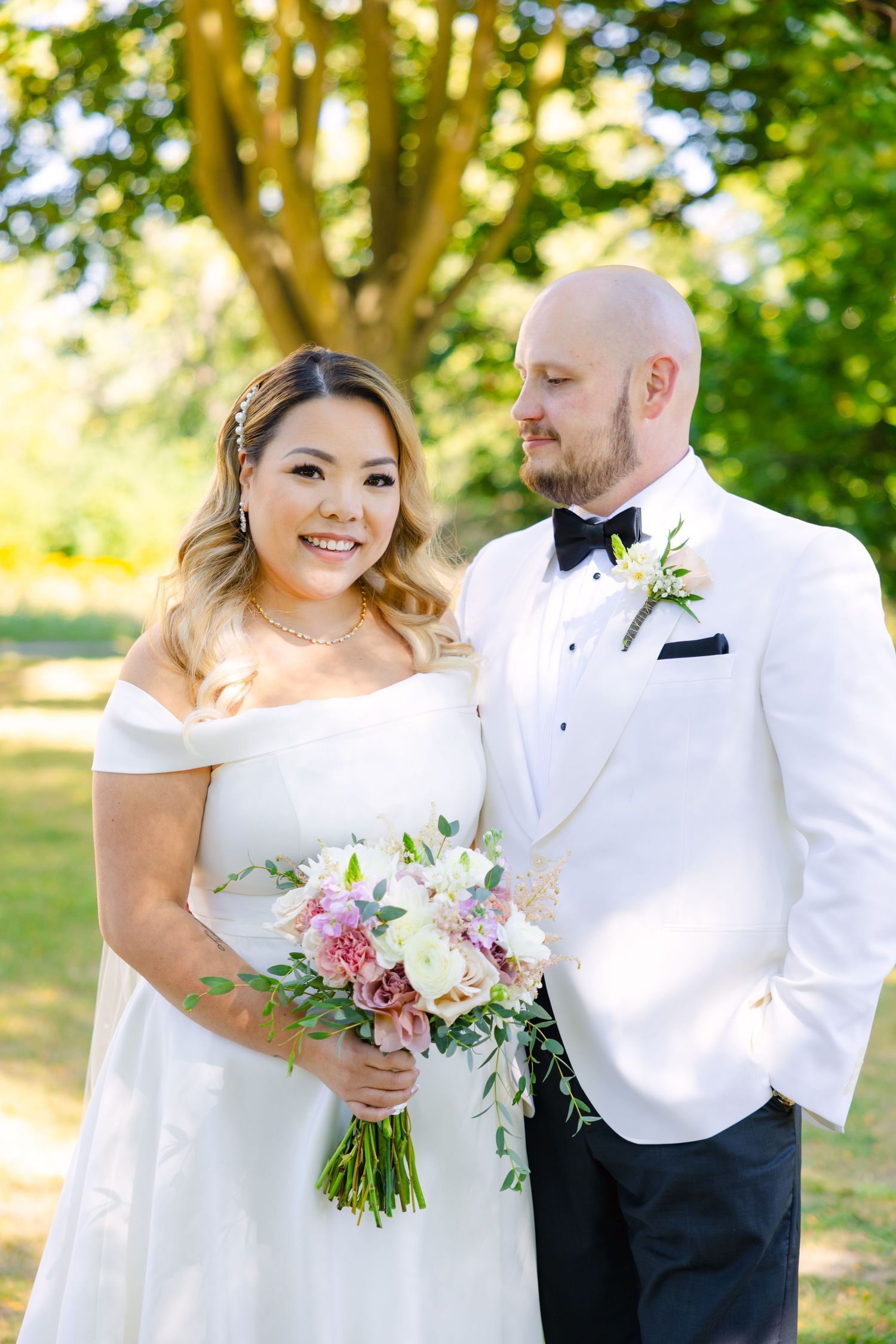 Intimate wedding portrait of couple in white attire against background of leafy trees.