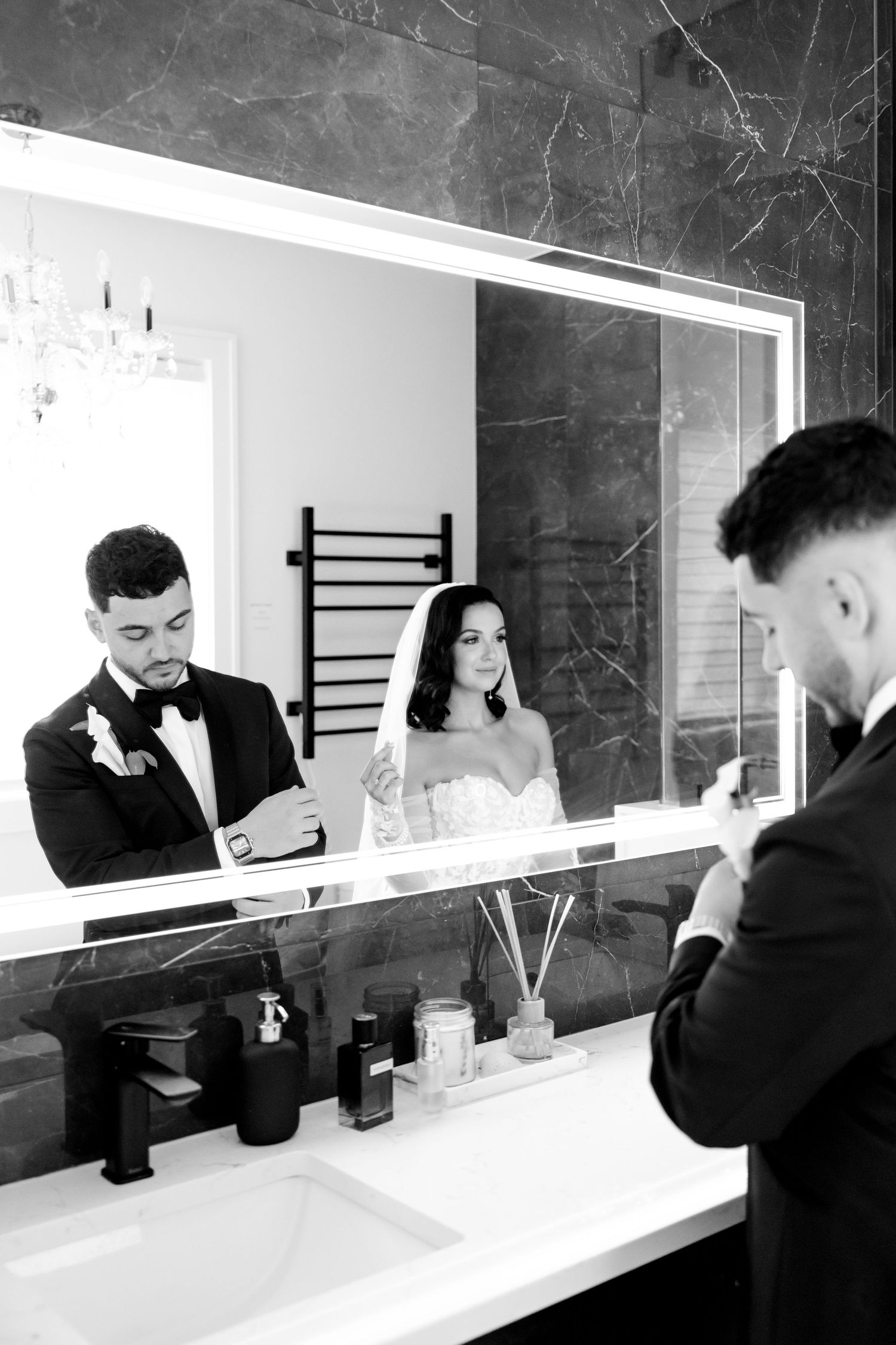 Wedding couple's reflection in bathroom mirror shows them adjusting formal attire.