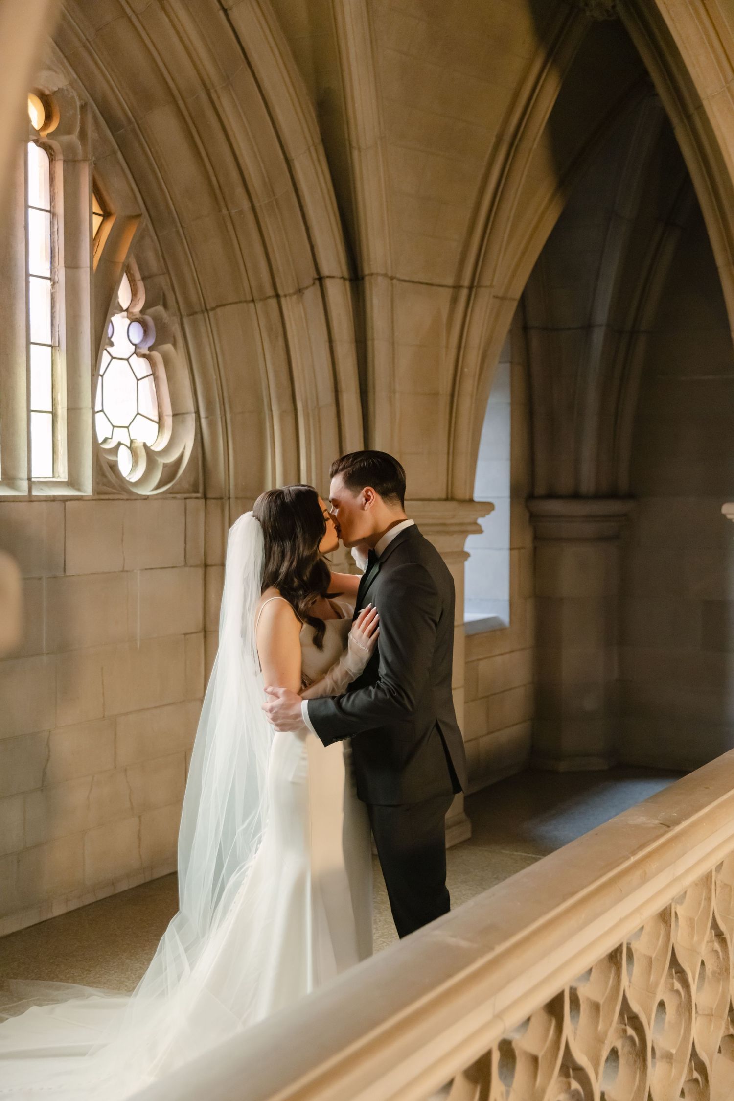 Newlyweds embrace in an elegant stone corridor with gothic architectural details and natural lighting.