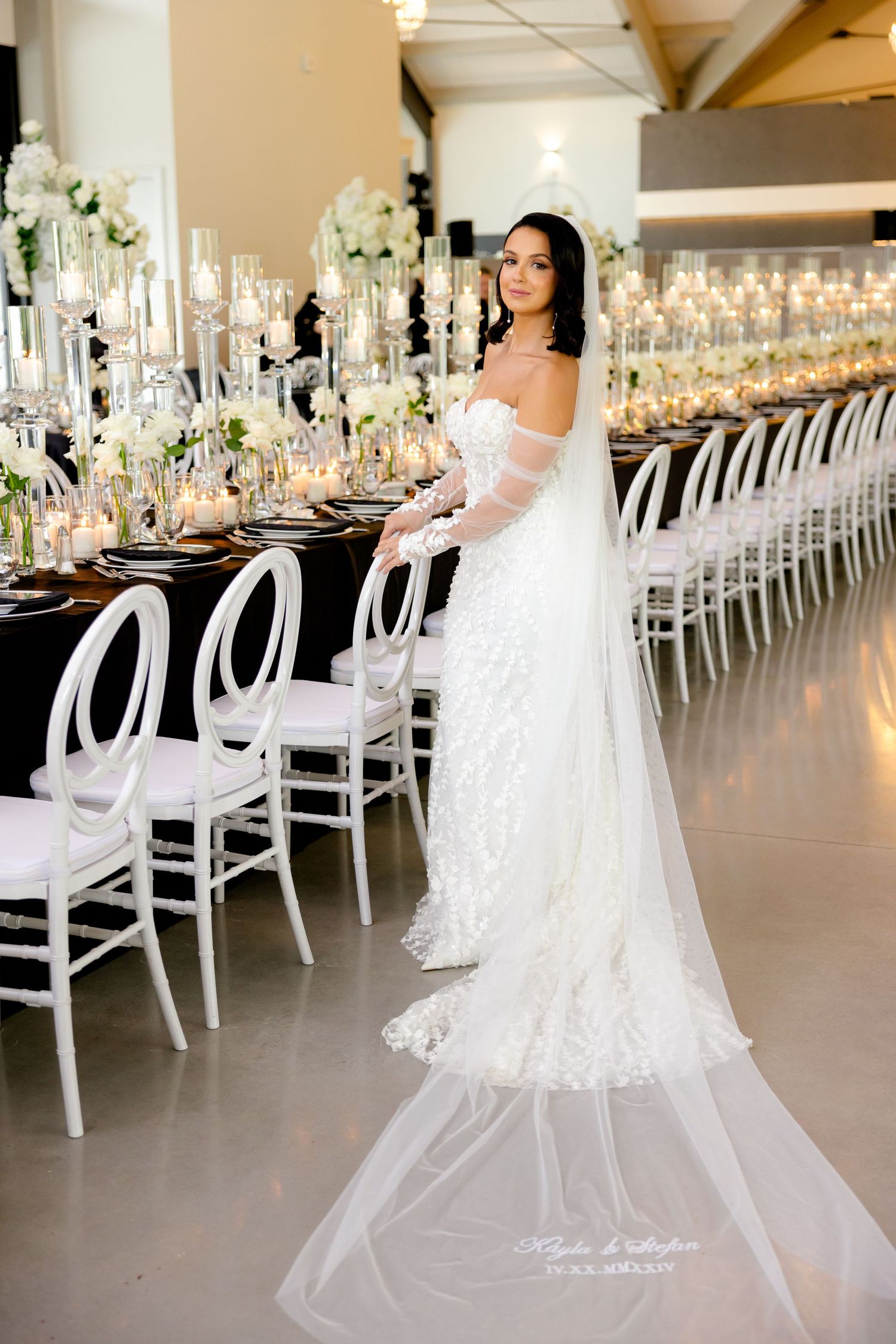 An elegant white wedding gown shown in a beautifully decorated reception hall with tall floral centerpieces and candles.