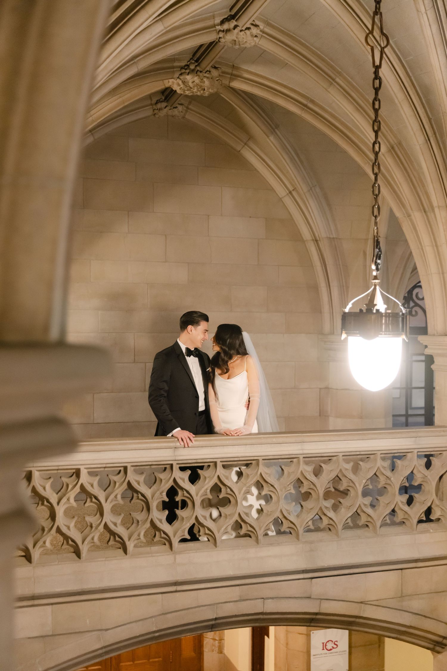 A romantic wedding moment captured under gothic arches with a decorative stone balustrade and hanging vintage lamp.