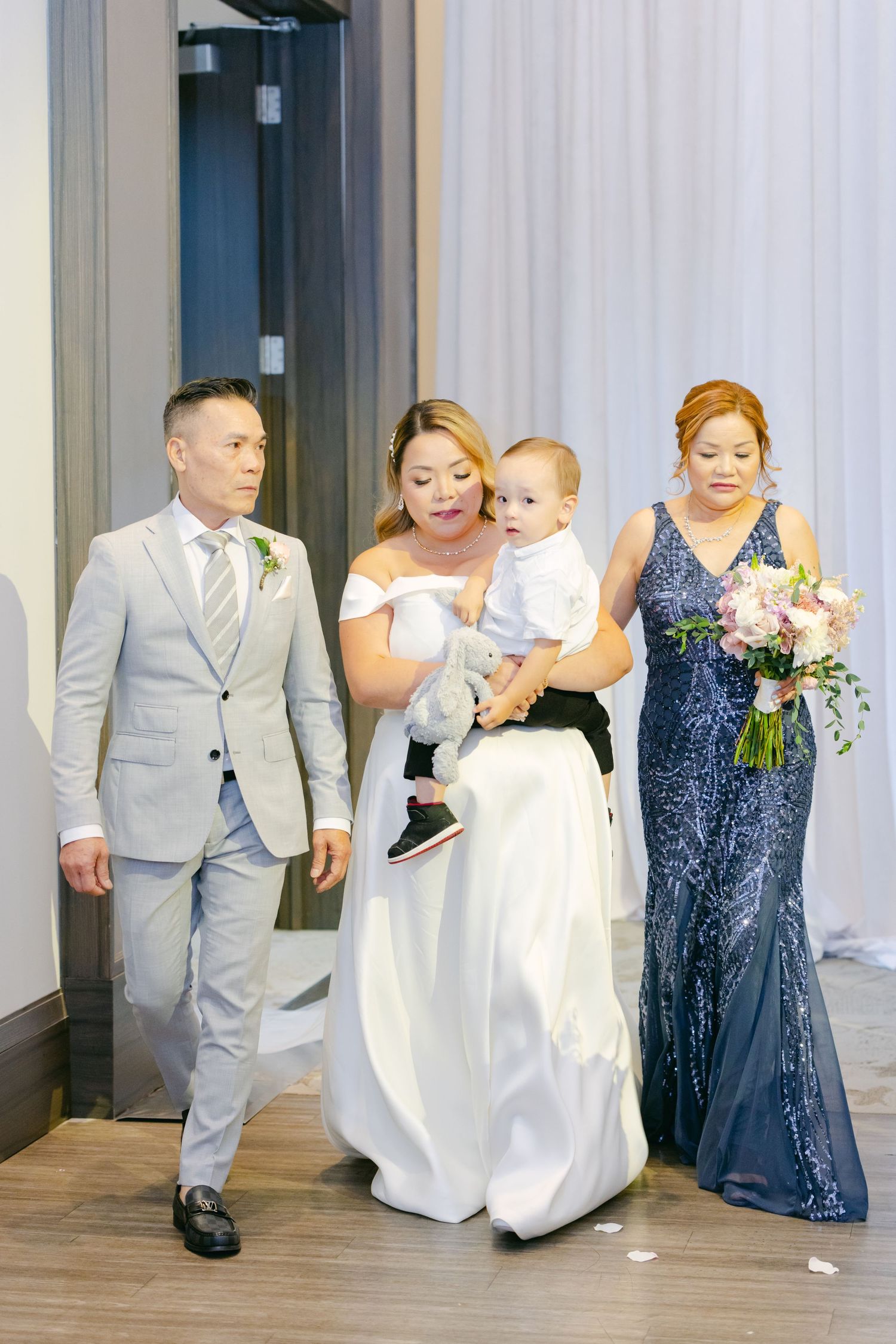 Wedding party members walking in formal attire with a baby at an indoor venue.