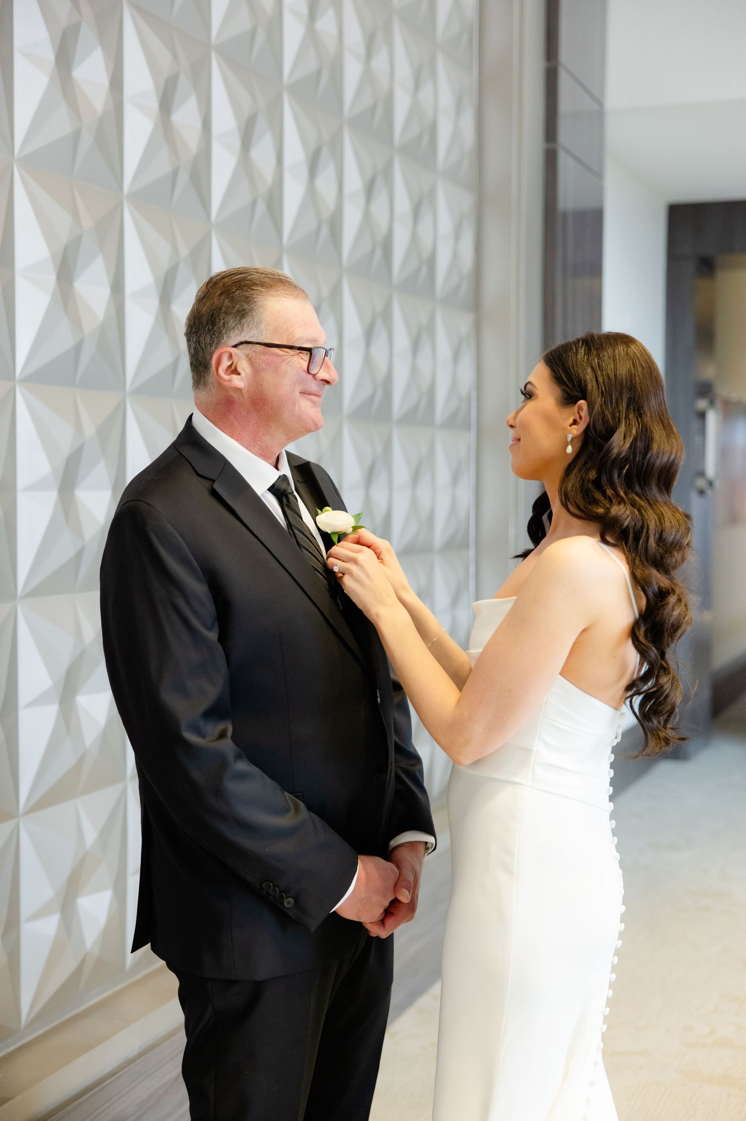 A bride fixes a boutonniere on a suit in front of a geometric textured wall.