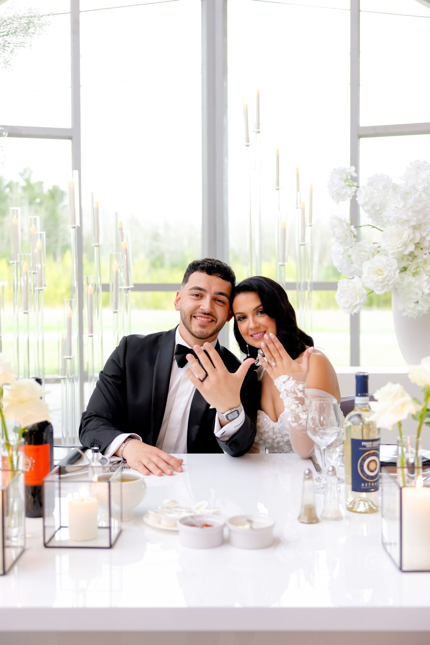 Newlyweds showing off their rings at a beautifully decorated sweetheart table with white floral arrangements.
