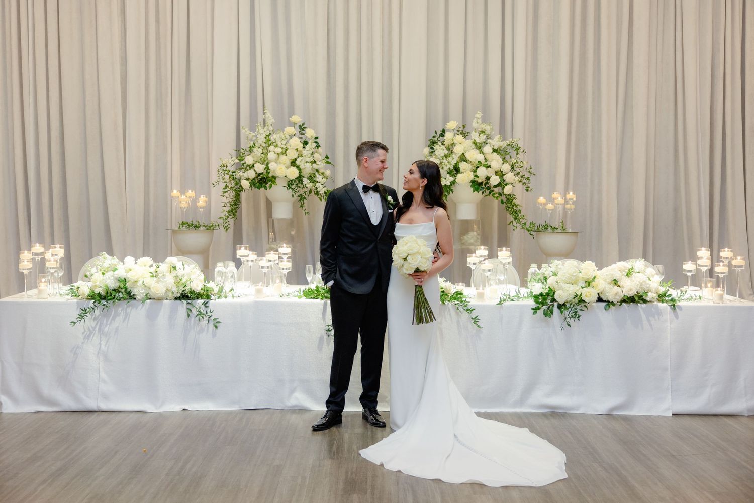Elegant wedding reception setup featuring white florals, draped backdrop and romantic lighting.