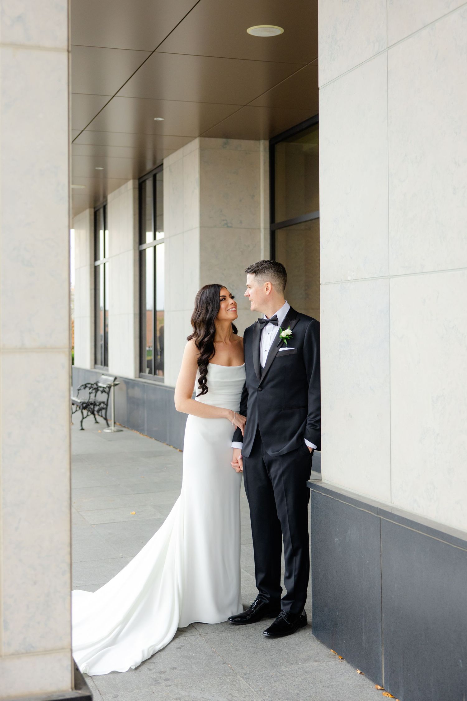 A newlywed couple poses elegantly against modern architecture in formal wedding attire.