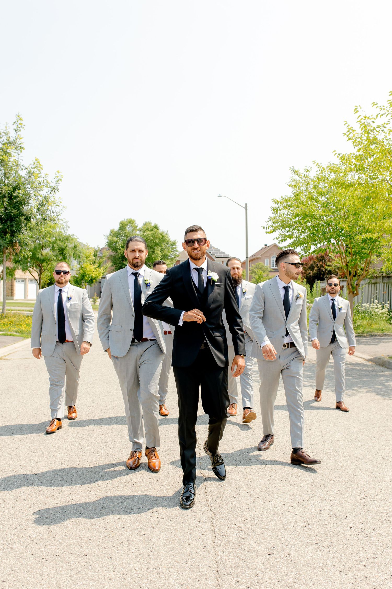 Groomsmen in light grey suits walking with central figure in black suit on residential street.