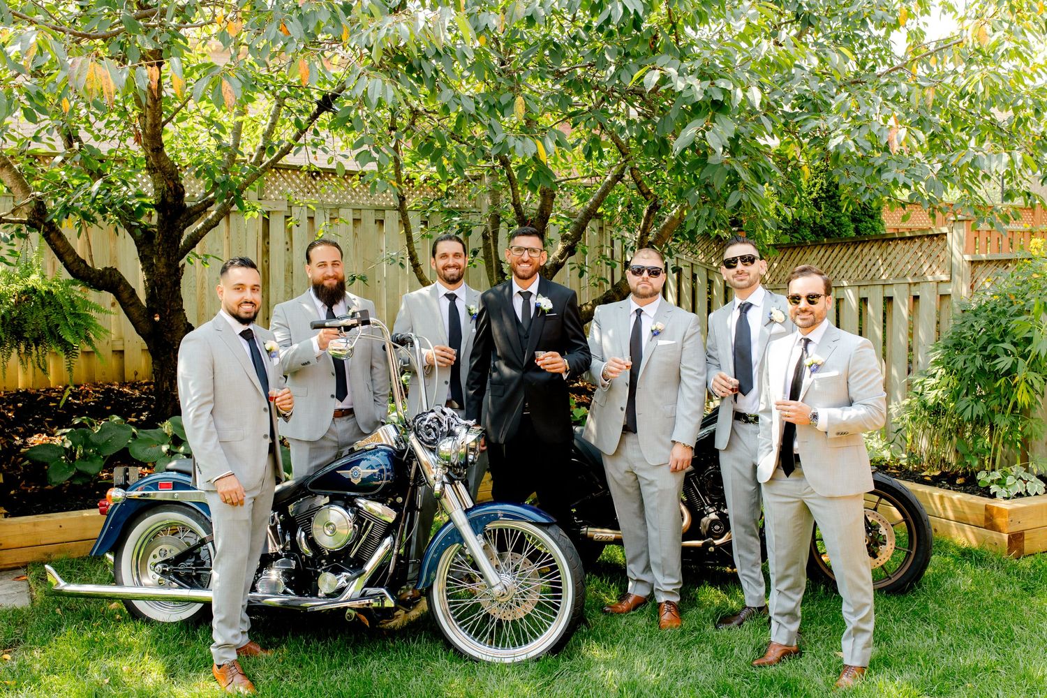 Wedding party in gray suits gathered around vintage motorcycles in a backyard garden.