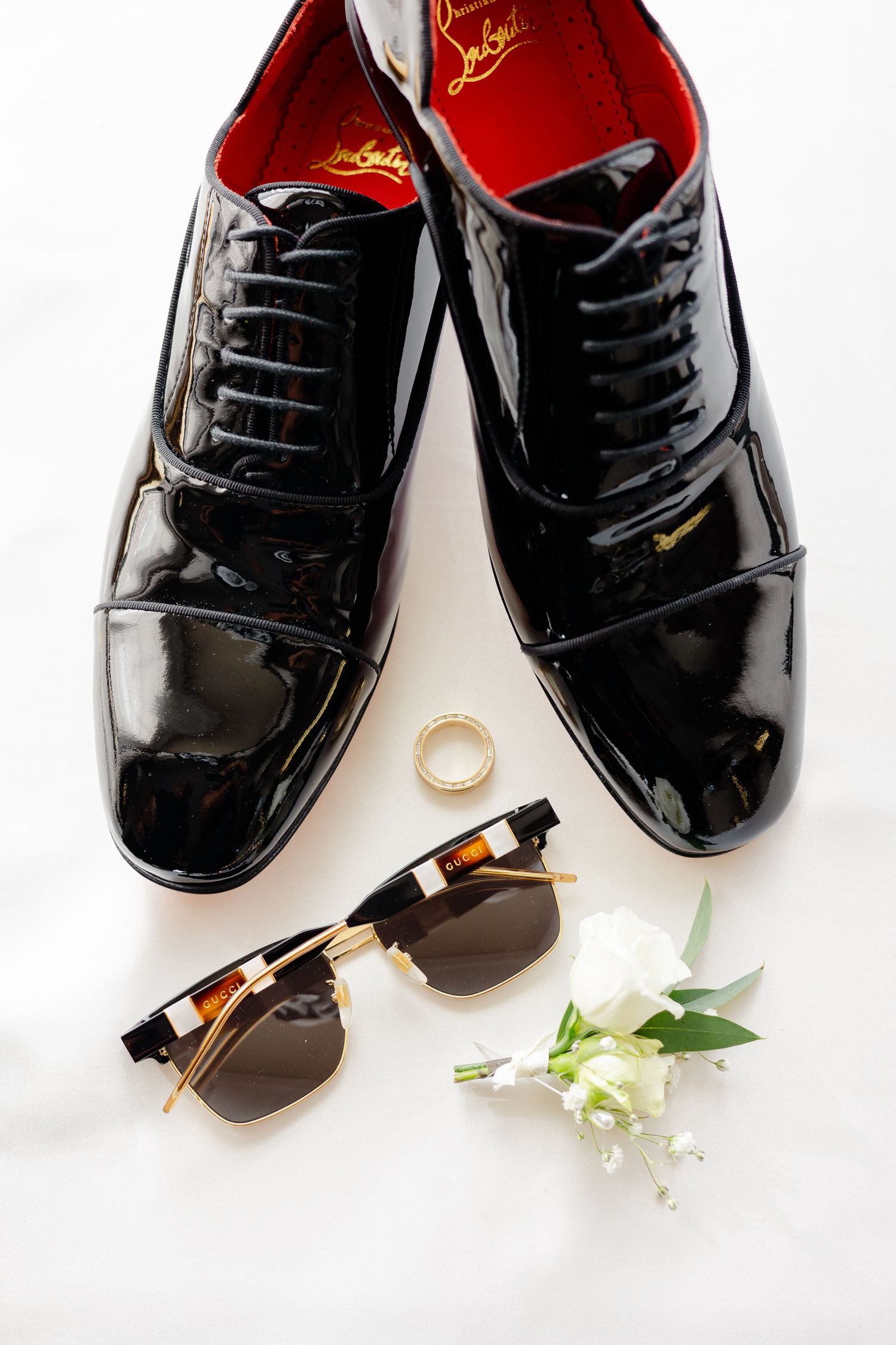 Shiny black patent leather dress shoes with sunglasses and wedding ring on white background.