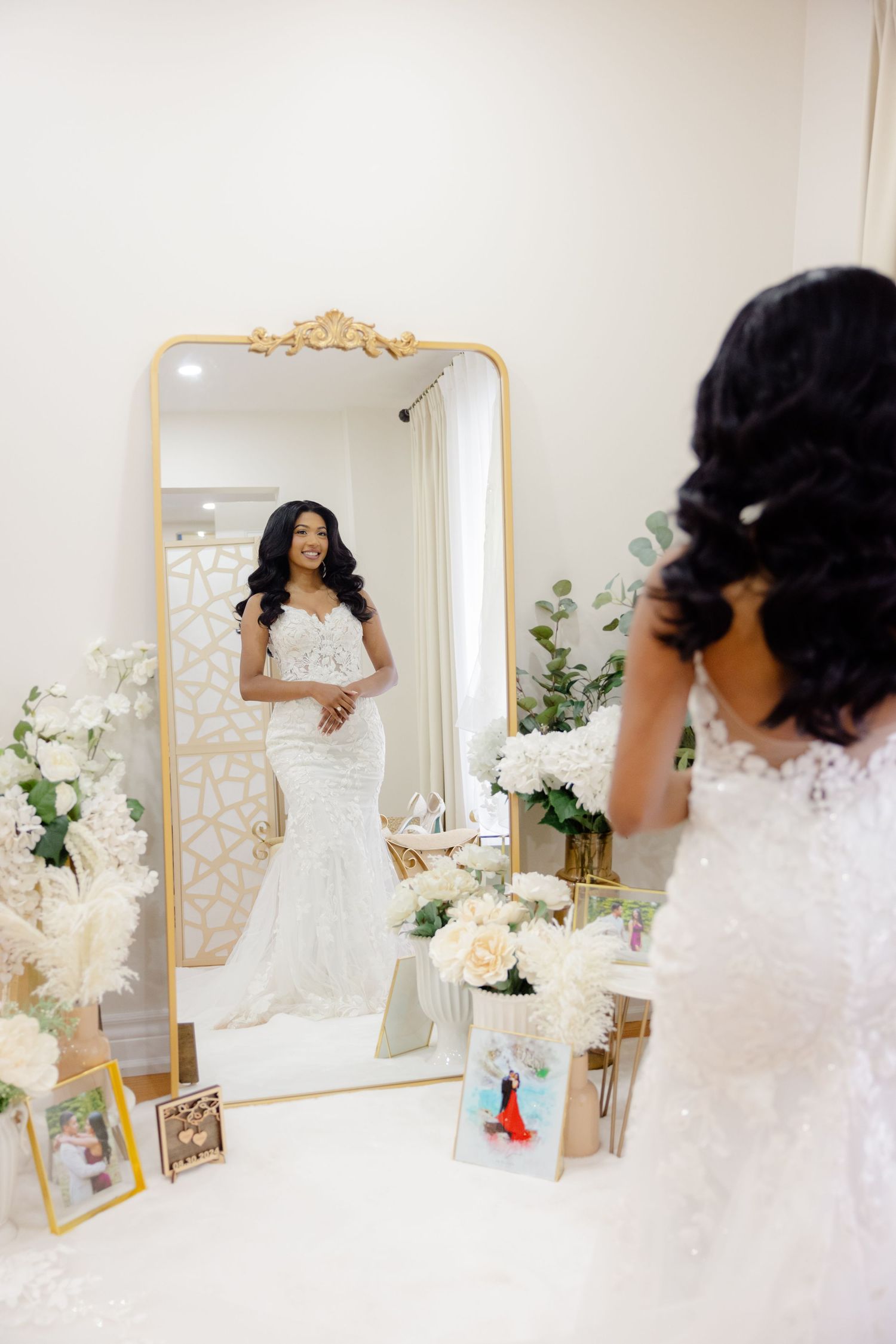 Wedding dress reflection in ornate gold mirror surrounded by white floral arrangements.